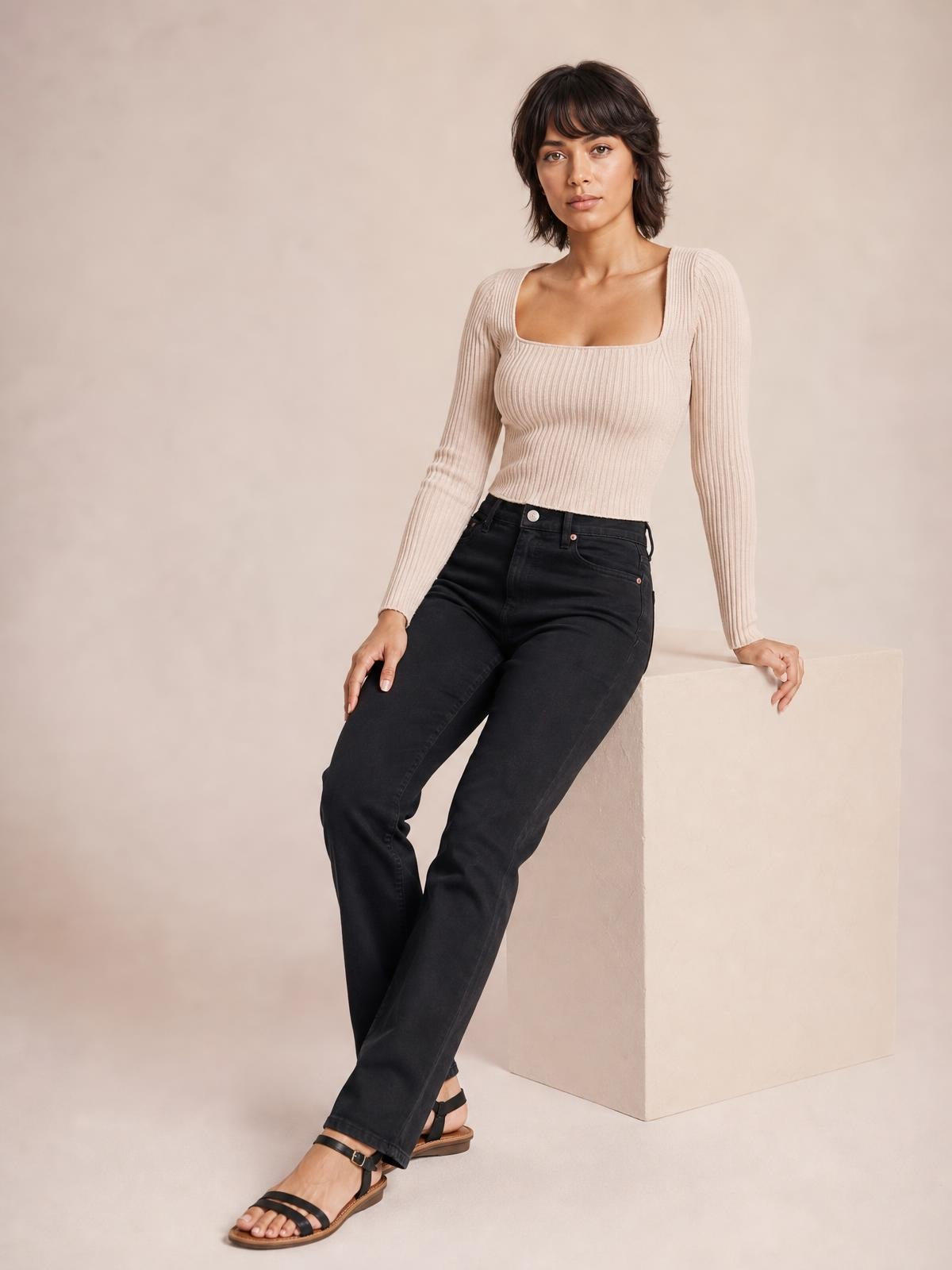 A model leans against a block wearing a light beige ribbed sweater with a square neckline, fitted silhouette, long sleeves, and cropped hem.