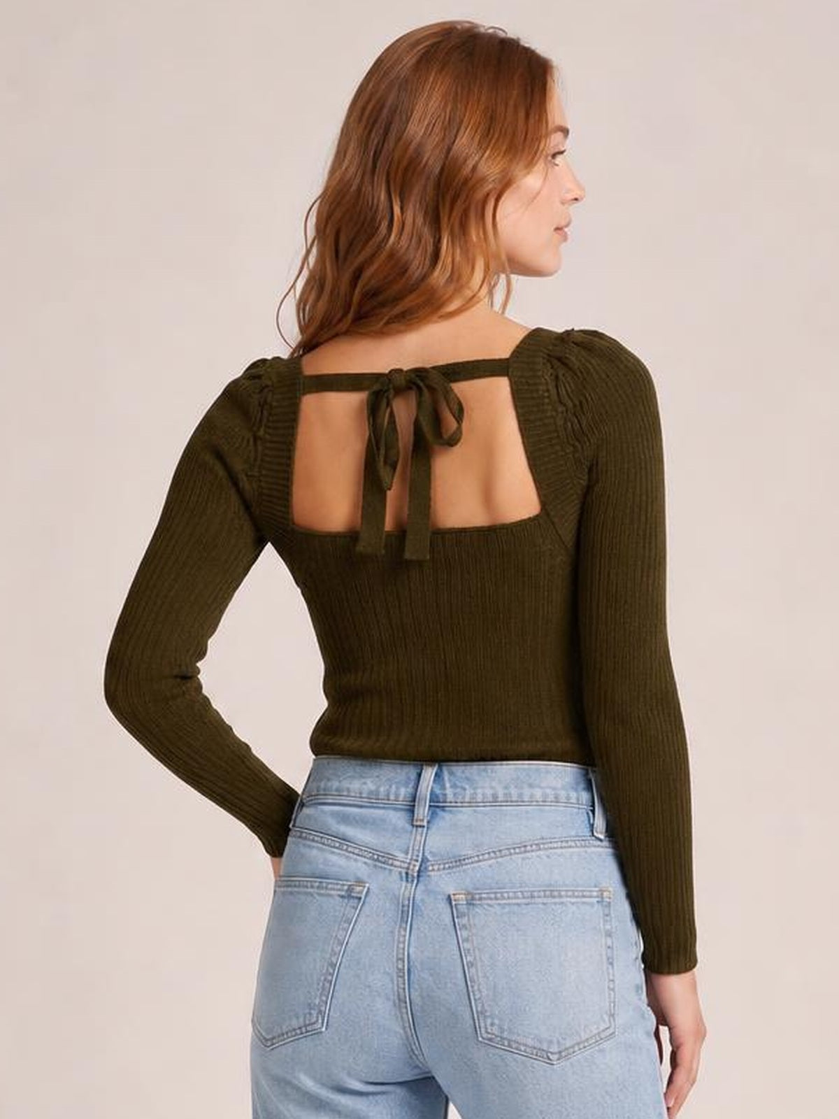 A model poses in profile wearing an olive ribbed long sleeve sweater with a fitted silhouette, square open back, and tied strap detail.
