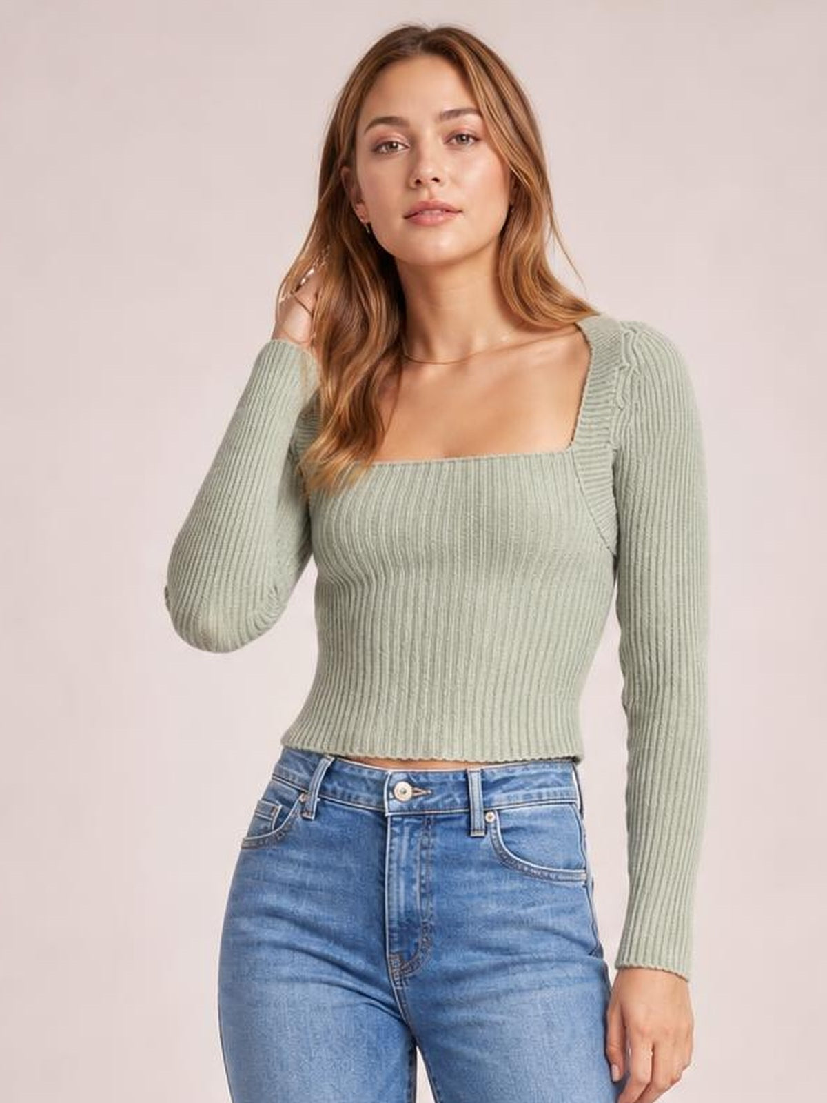 Model poses with one hand in her hair wearing a pale sage ribbed square neck long sleeve sweater with a fitted, waist-length silhouette.