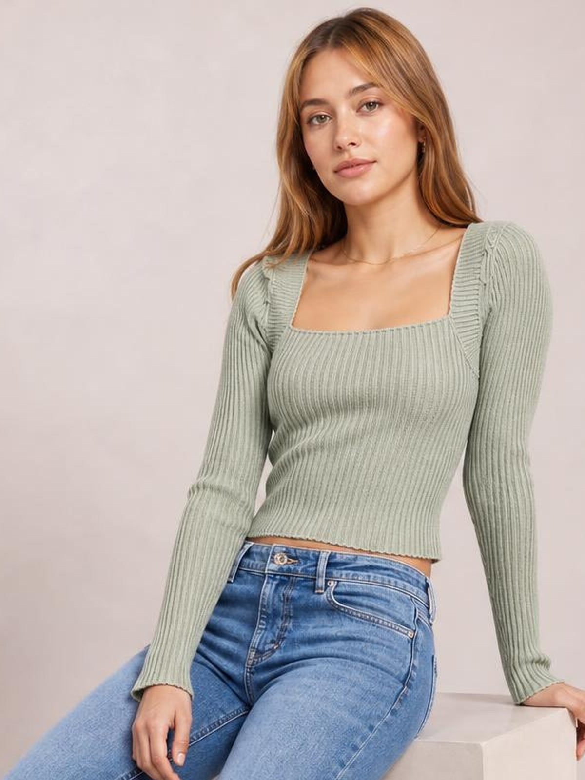 Model seated in a sage green ribbed square neck long sleeve sweater with a fitted cropped silhouette and stretchy knit construction.