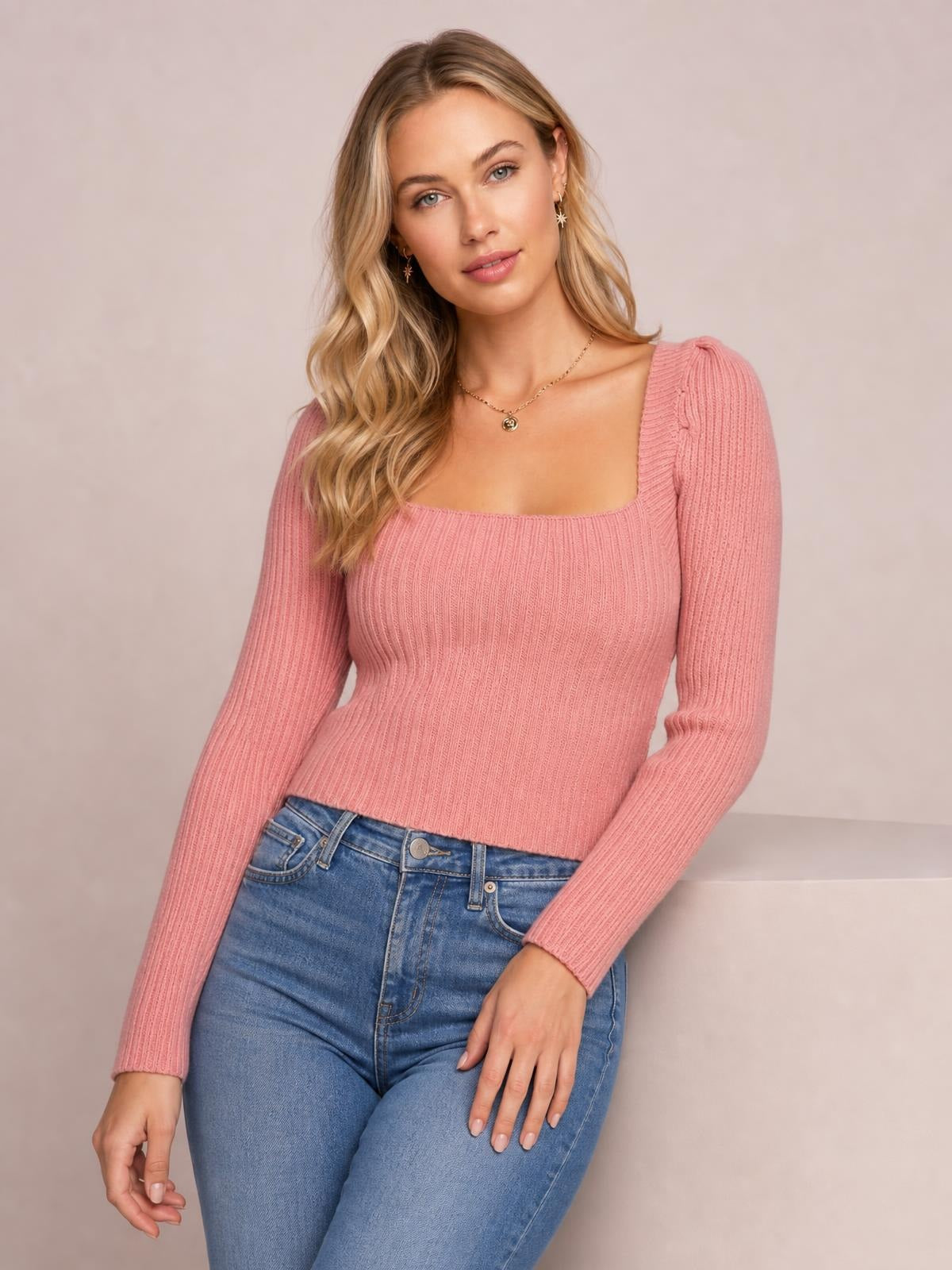 Model poses in a pink ribbed sweater with a square neckline, long fitted sleeves, a close body-hugging silhouette, and a cropped hem.