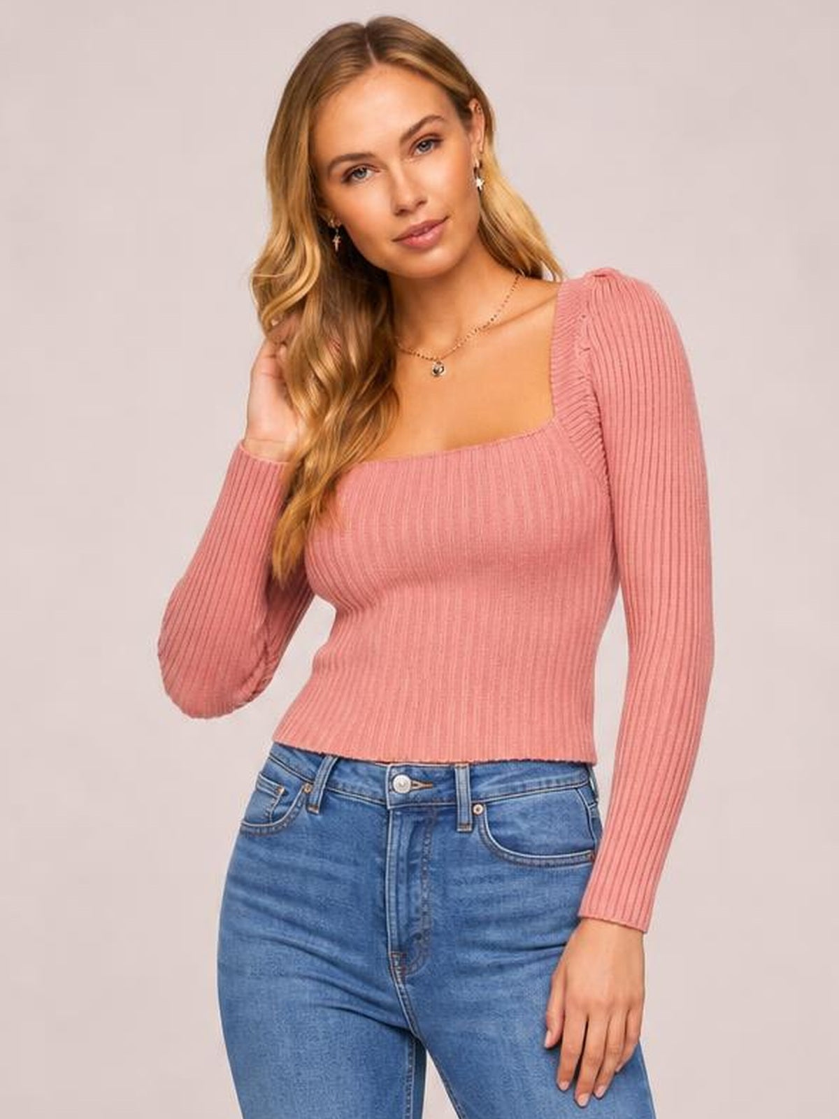 Model poses in a pink ribbed square neck long sleeve sweater with a fitted silhouette, gently holding her wavy hair.