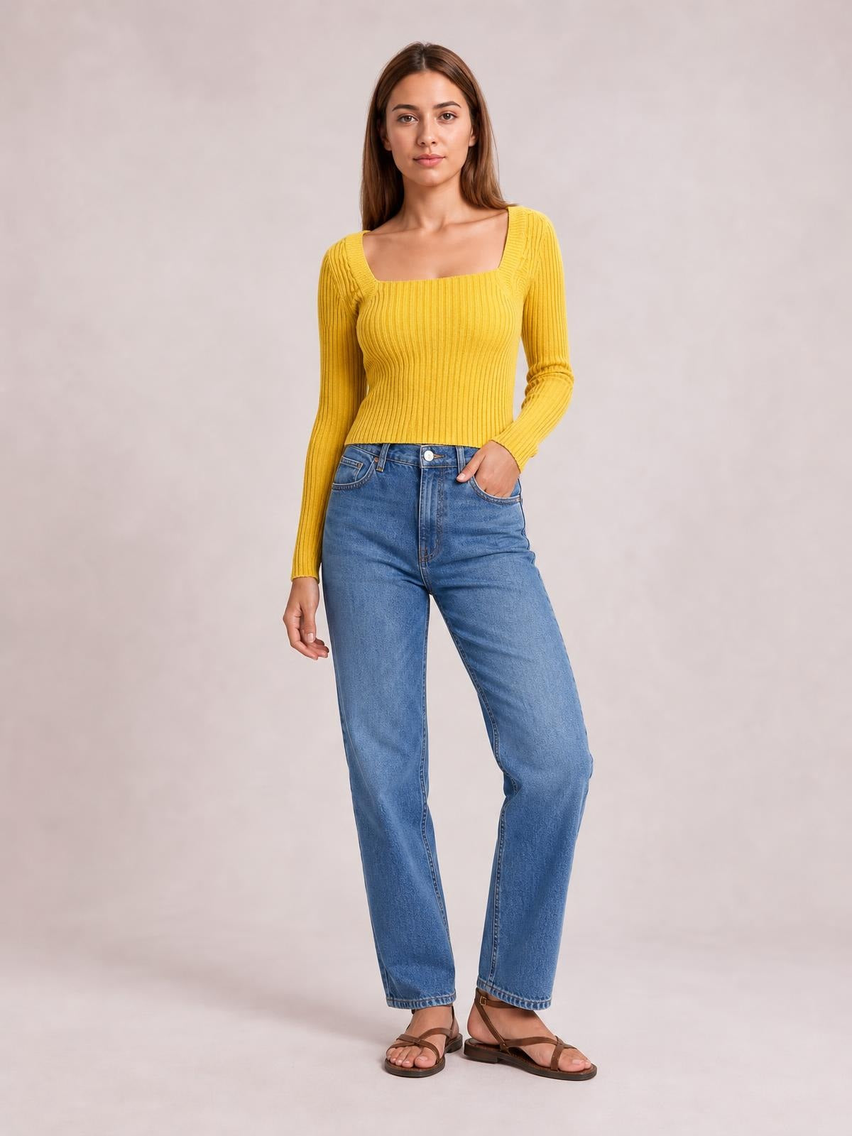 Model wears a mustard yellow ribbed square neck long sleeve sweater with a fitted silhouette and one hand tucked into her jeans pocket.
