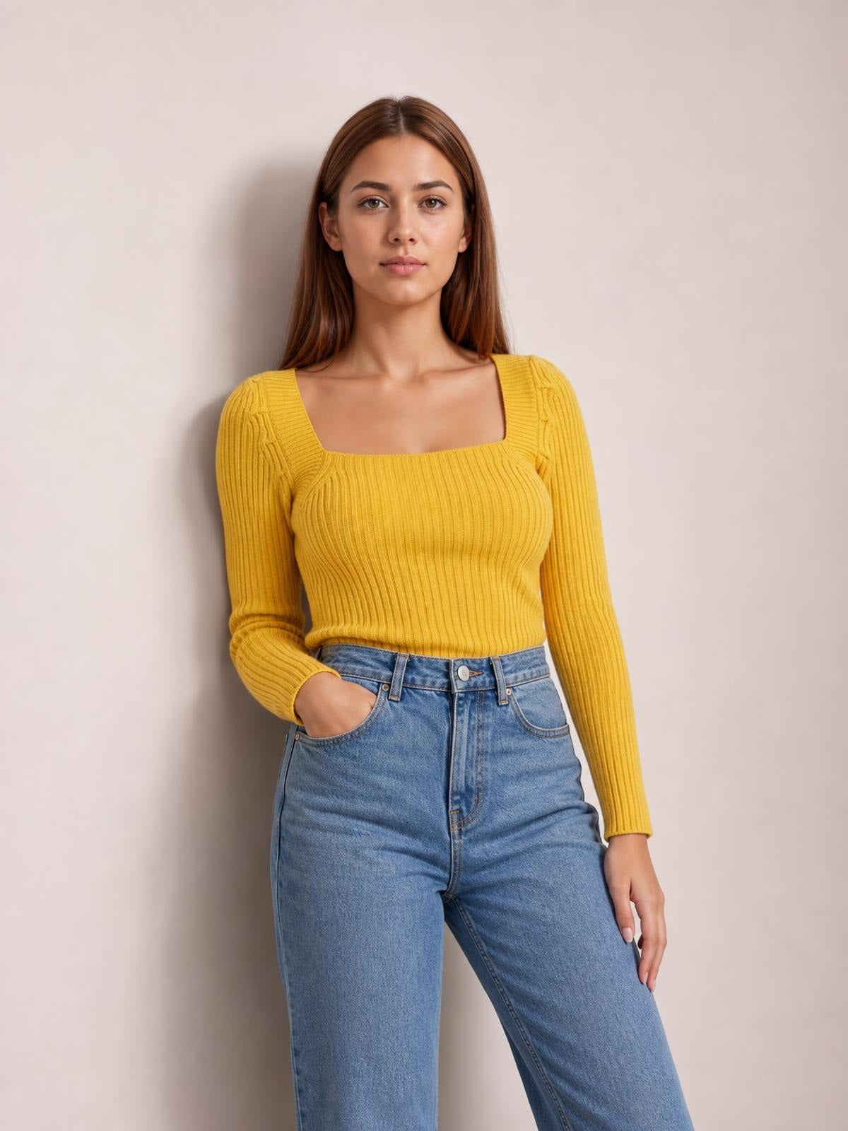A model poses in a fitted mustard-yellow ribbed sweater with a square neckline and long sleeves, tucked into high-waisted blue jeans.