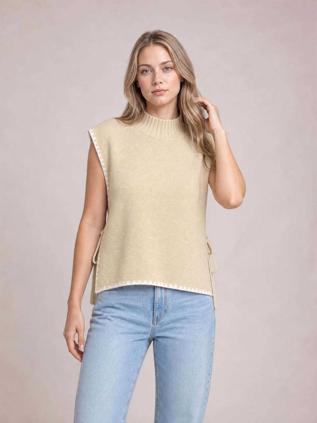 Model wearing a cream ribbed turtleneck sweater vest with contrast trim, sleeveless straight silhouette, side ties, and a relaxed fit, posing with one hand in her hair.