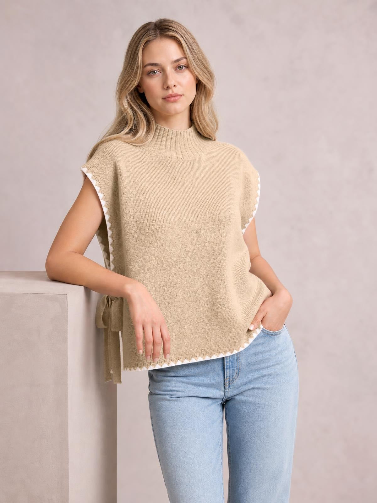 Model wearing a beige ribbed turtleneck sweater vest with contrast white trim, relaxed sleeveless silhouette, side ties, and a scalloped hem.