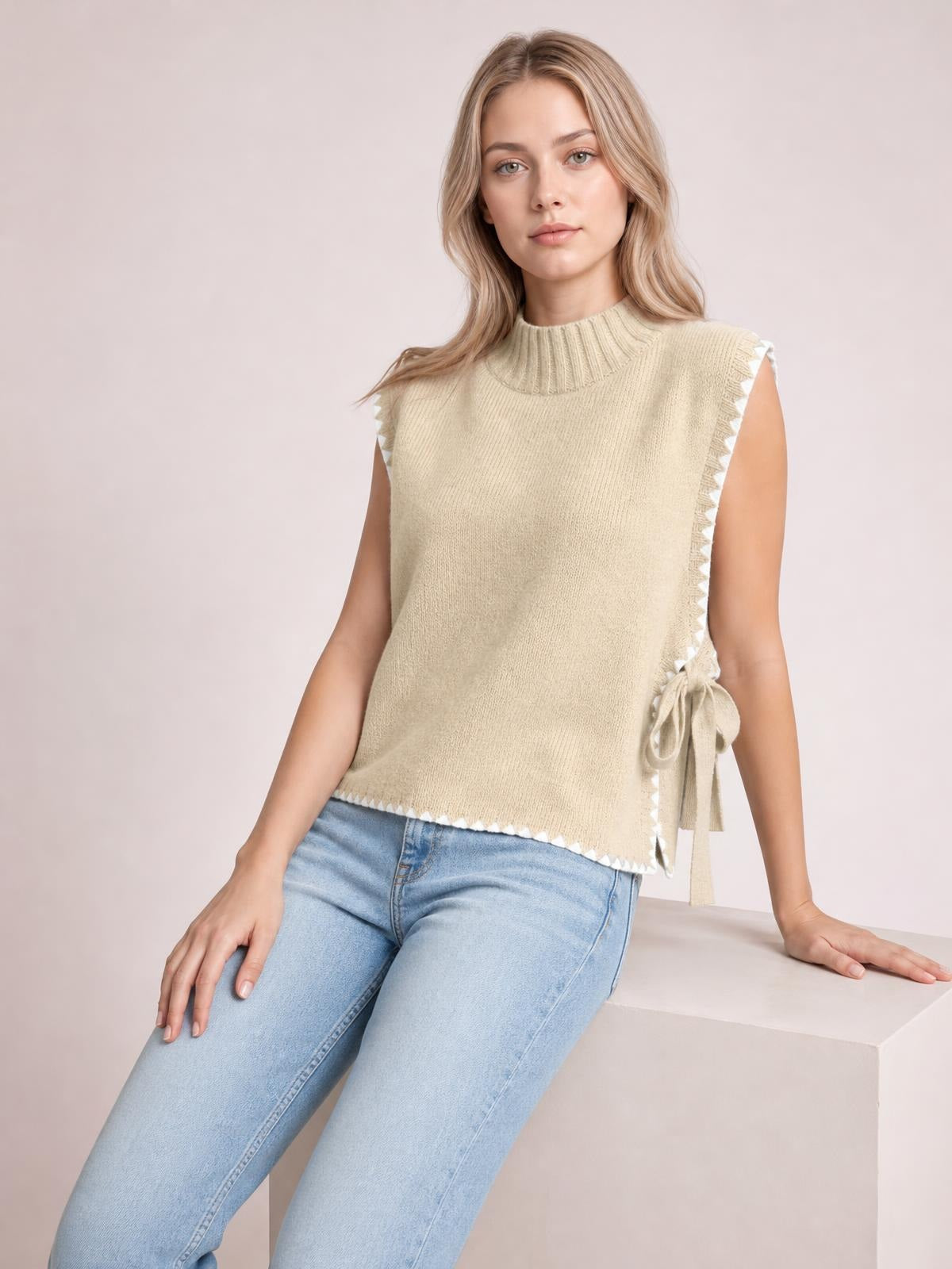 A model leans against a ledge wearing a beige ribbed turtleneck sweater vest with contrast trim, a relaxed silhouette, and side ties.