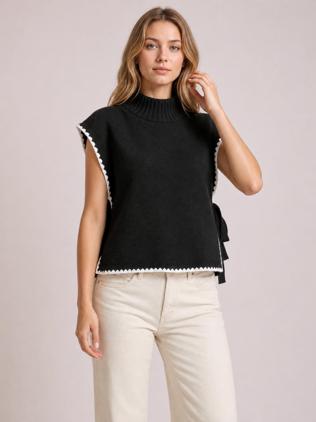 A model poses in a black ribbed turtleneck sweater vest with white contrast trim, relaxed straight silhouette, and side-tie details.