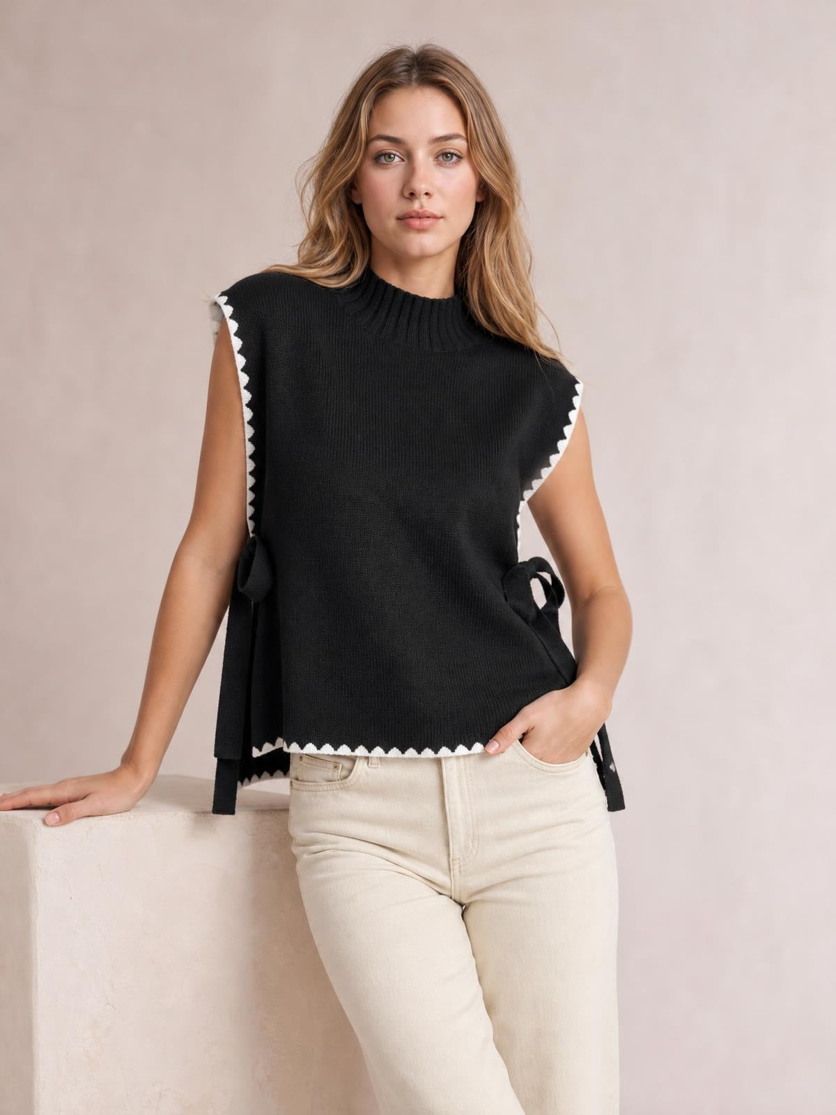 Model wears a black ribbed turtleneck sweater vest with white contrast trim, sleeveless silhouette, scalloped edges, and tied side openings.