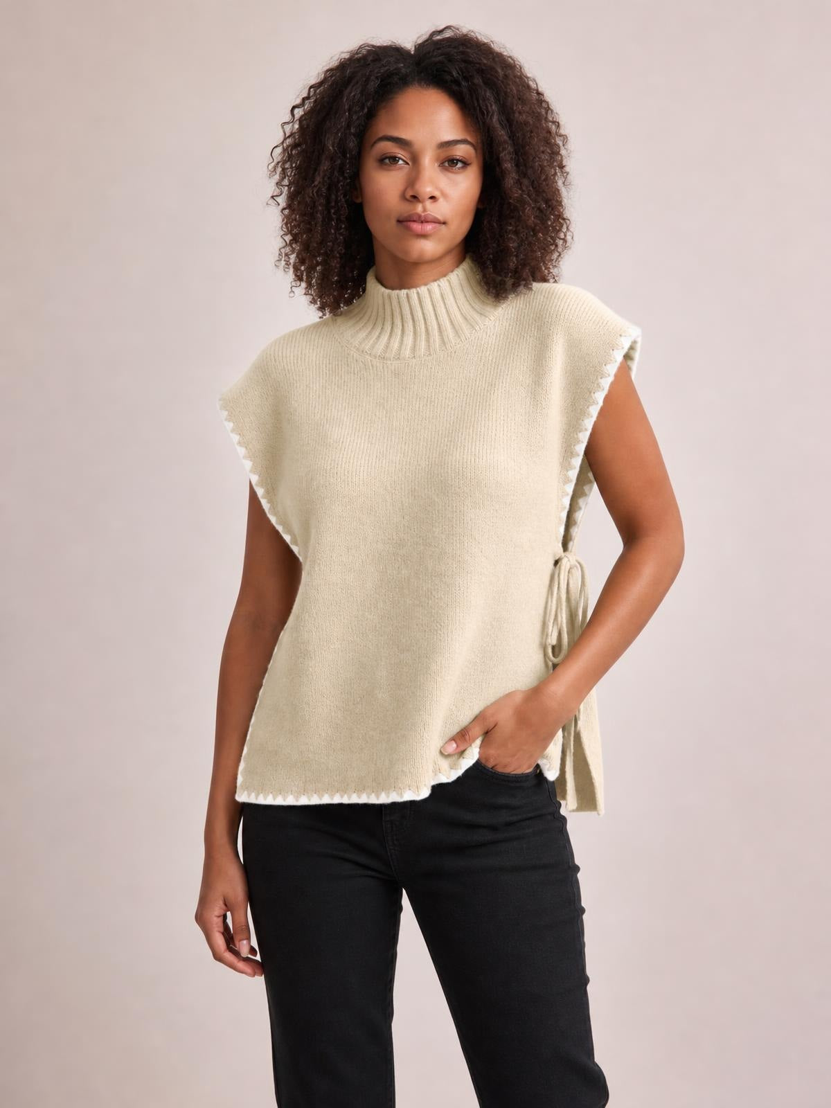 Model poses in a cream ribbed turtleneck sweater vest with contrast trim, side tie detail, relaxed sleeveless silhouette, and scalloped edges.