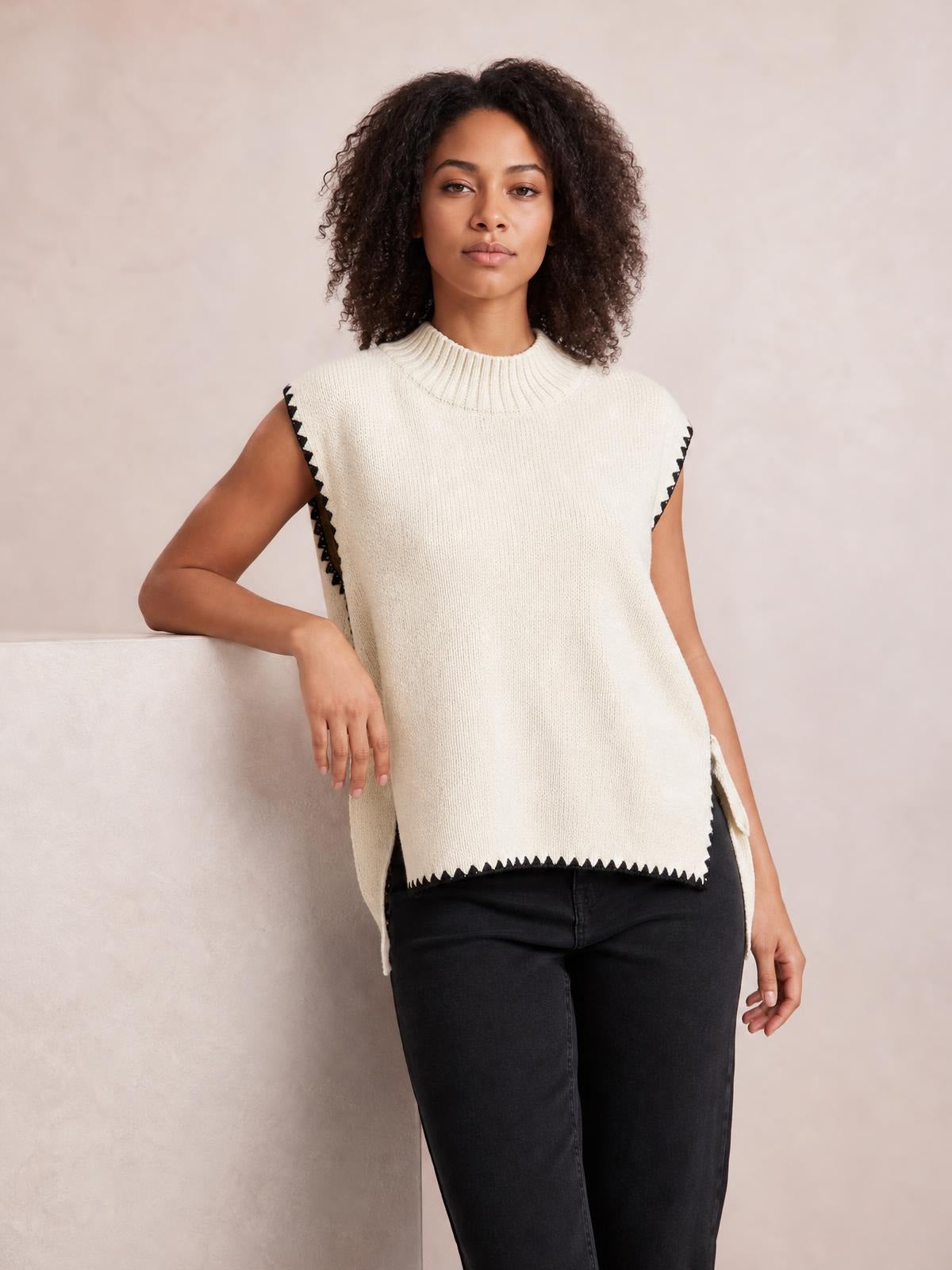 Model wears an ivory ribbed turtleneck sweater vest with black contrast trim, side slits, and a relaxed long silhouette while leaning on a ledge.