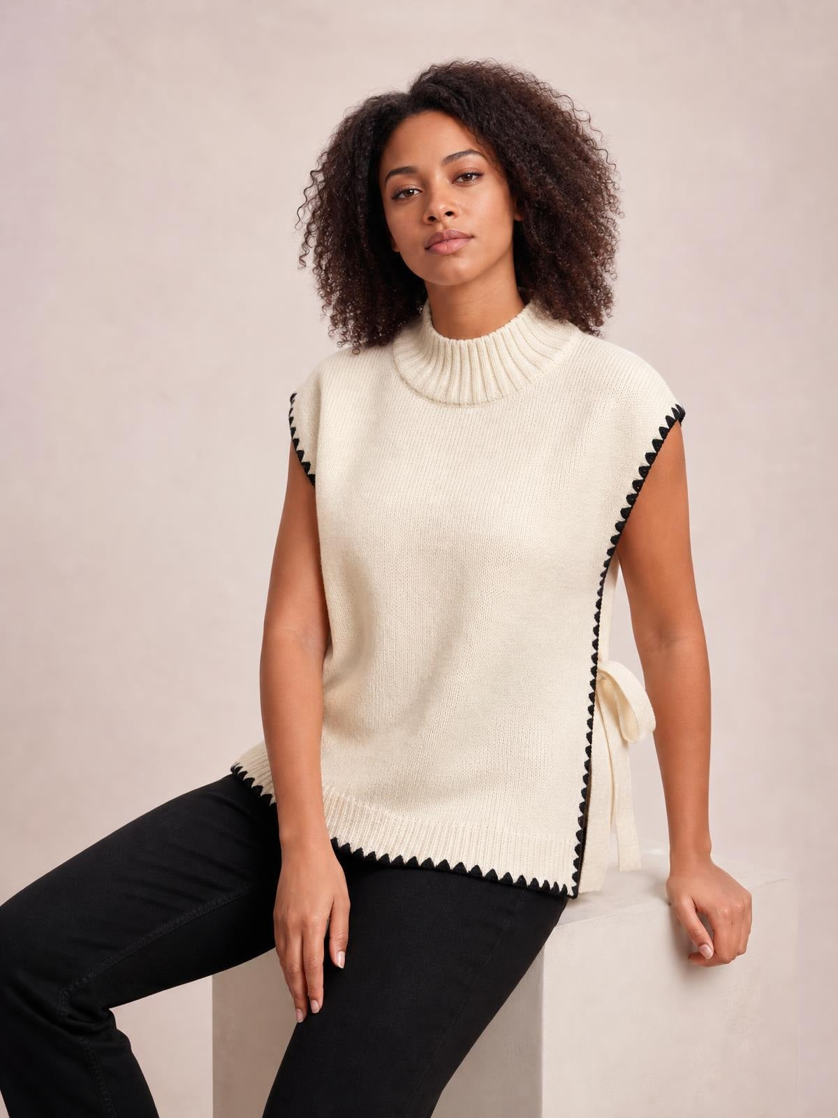 Model seated in a cream ribbed turtleneck sweater vest with black contrast trim, relaxed sleeveless silhouette, side tie, and scalloped hem.