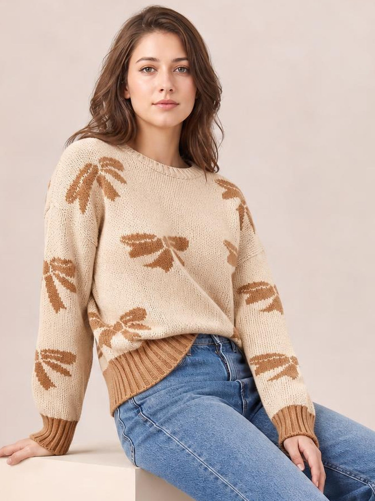 A woman seated in a cream drop-shoulder sweater with brown ribbon bow motifs, relaxed silhouette, and ribbed cuffs and hem.