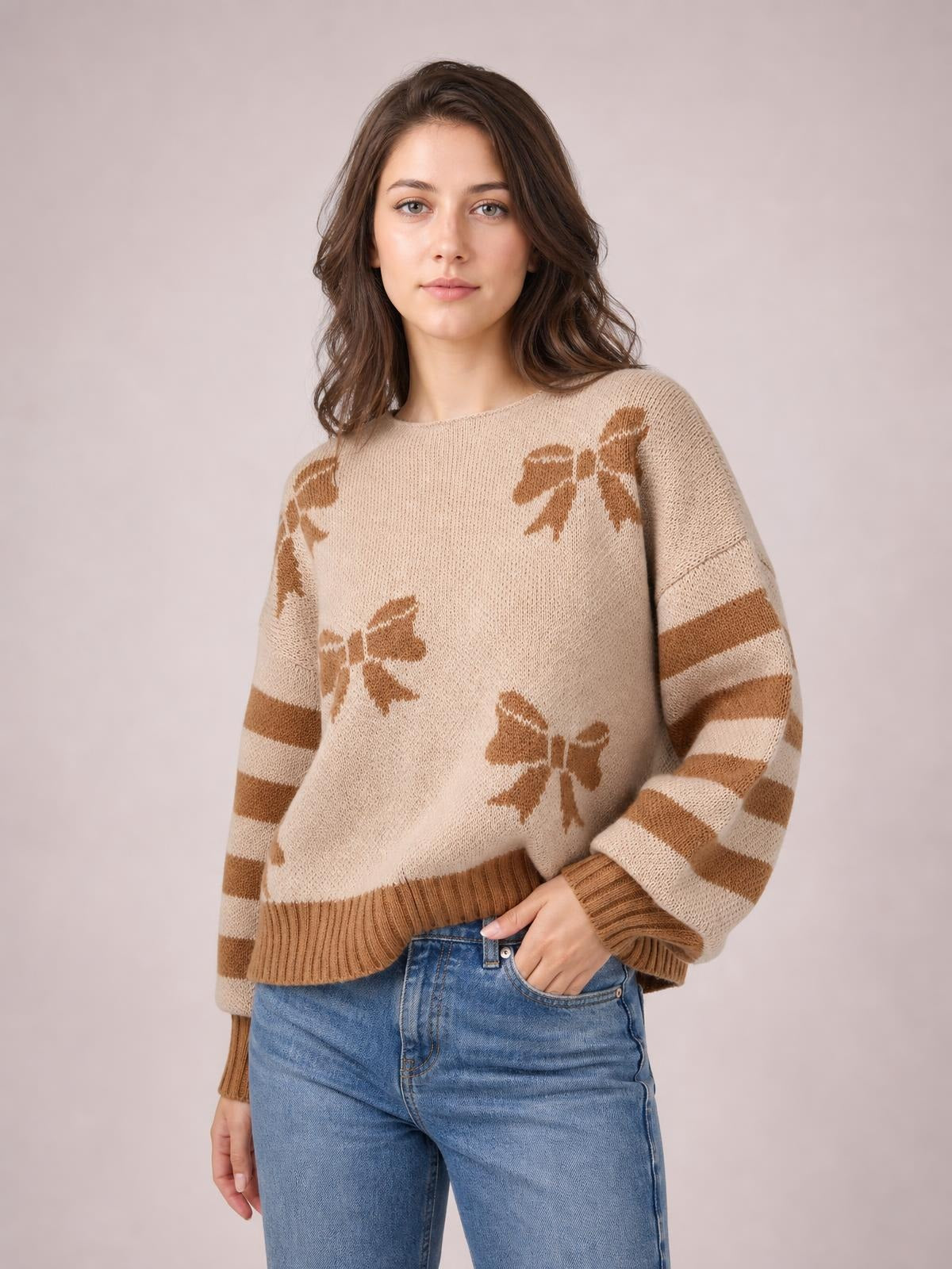 Model wearing a beige drop-shoulder sweater with brown ribbon bow motifs, striped sleeves, a relaxed silhouette, and ribbed neckline, cuffs, and hem.