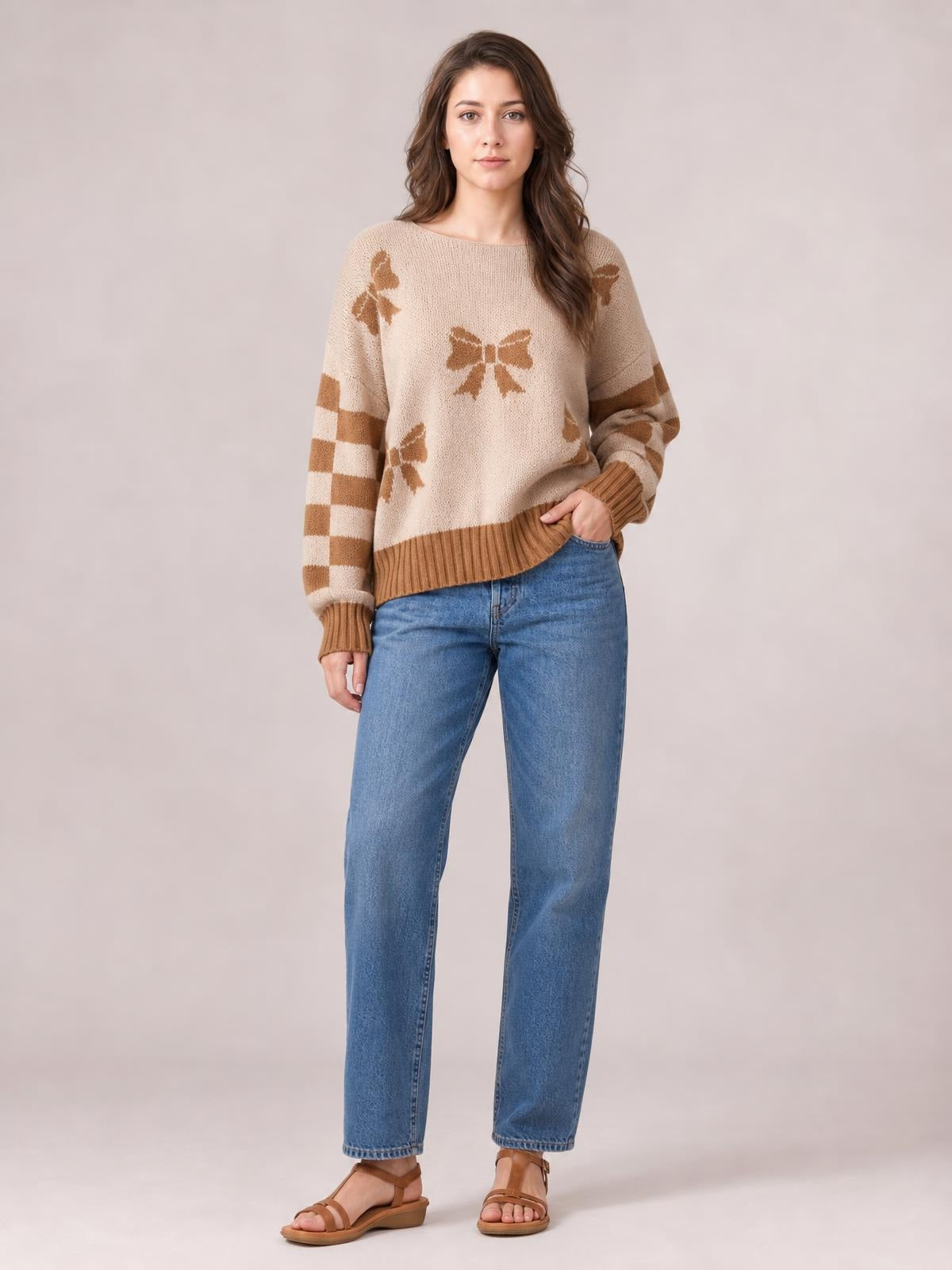 A woman models a cream drop-shoulder sweater with tan ribbon bow motifs, checkered striped sleeves, and ribbed cuffs and hem, standing with one hand in her jeans pocket.