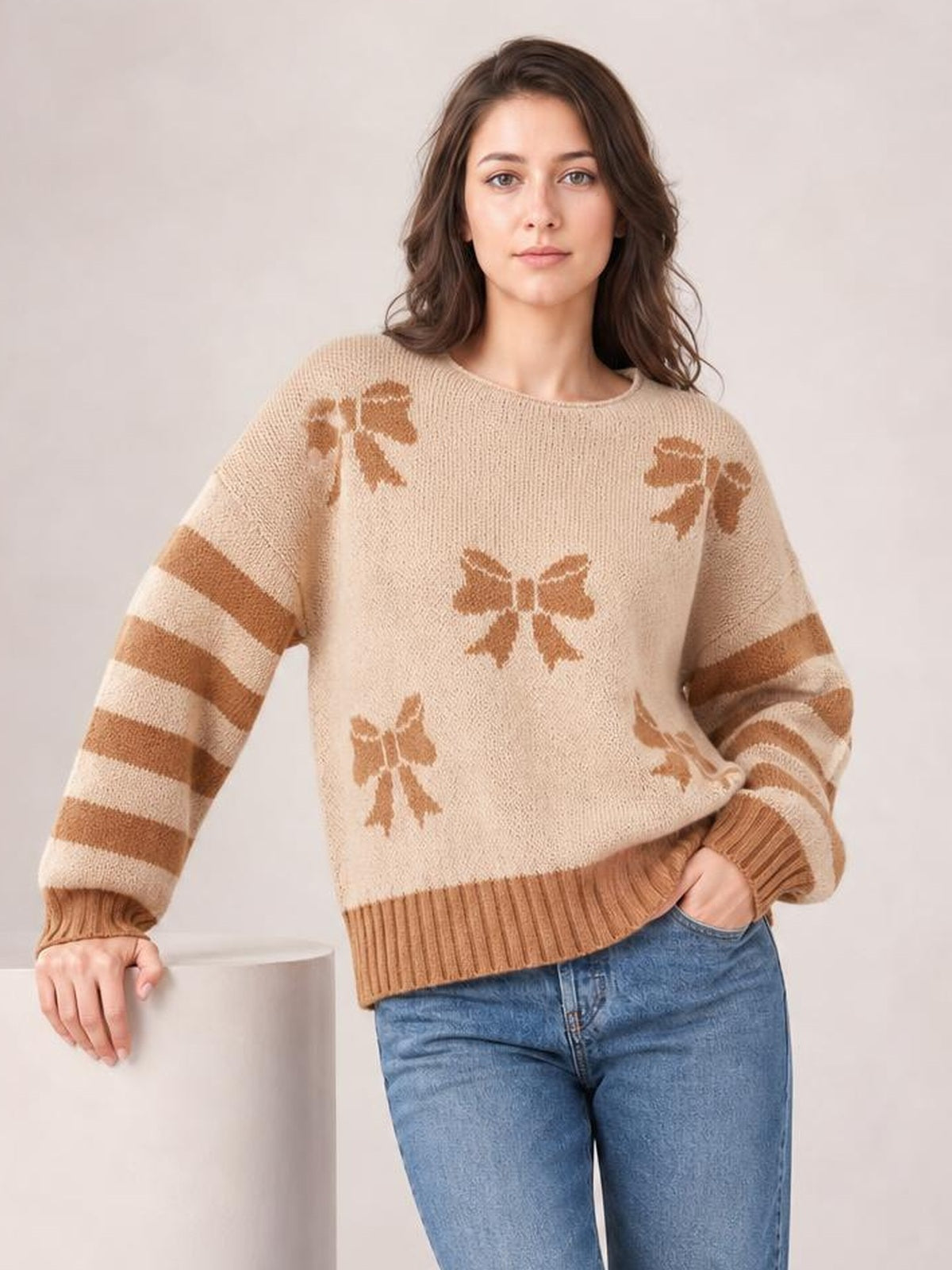 Model wears a beige drop-shoulder sweater with brown ribbon bow motifs, striped sleeves, and ribbed cuffs and hem in a relaxed silhouette.