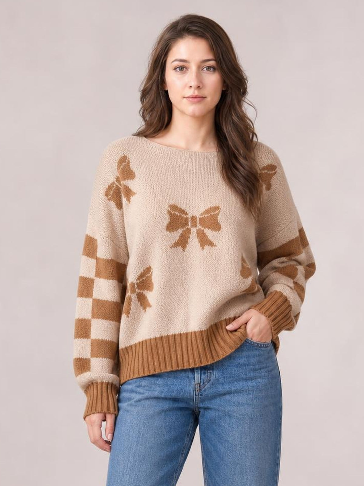 Model wearing a beige drop-shoulder sweater with brown ribbon bow motifs, checkered and striped sleeves, and ribbed cuffs and hem.