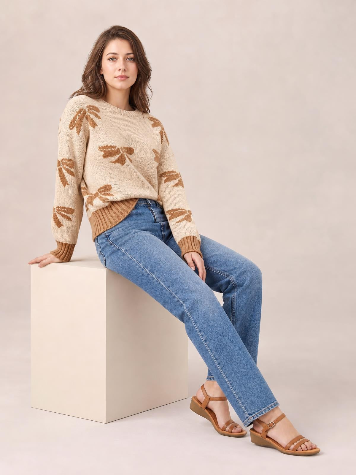 A seated woman wears a relaxed cream drop-shoulder sweater with brown ribbon bow motifs, ribbed crewneck, cuffs, and hem.