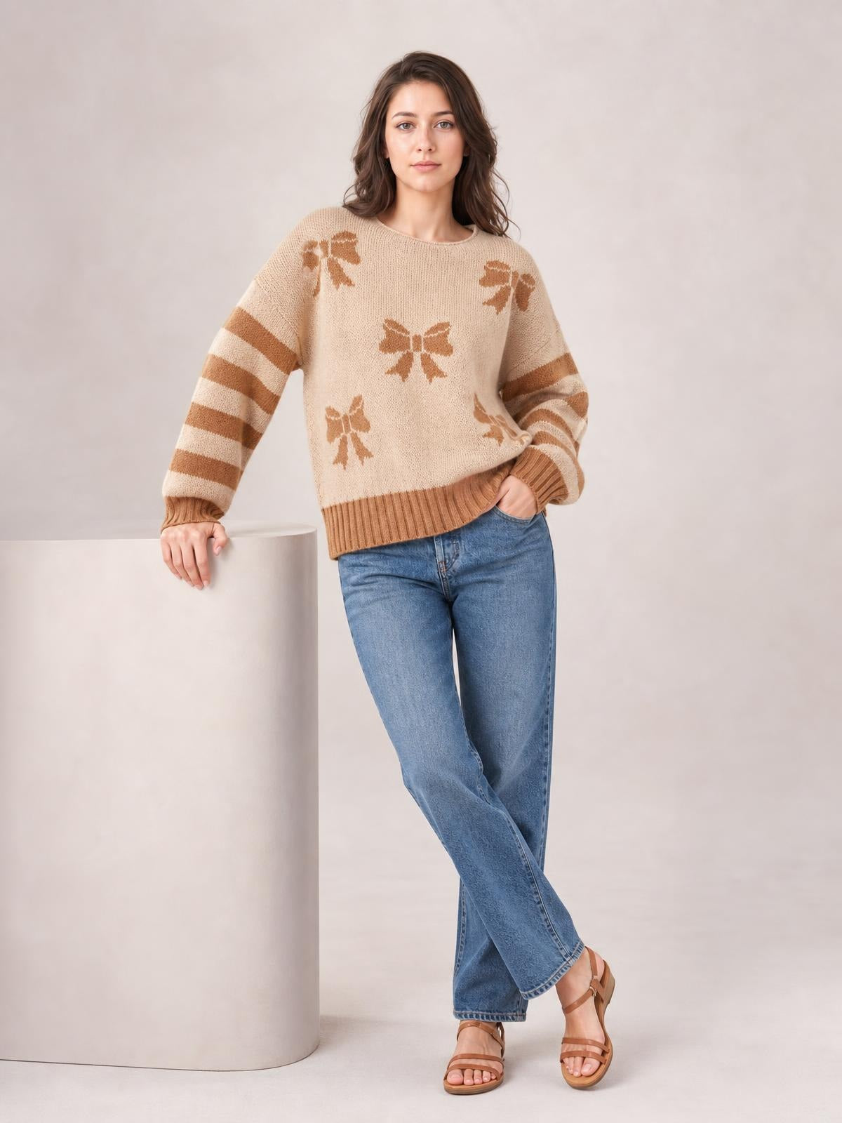 A model wears a beige drop-shoulder sweater with brown ribbon bow motifs, striped sleeves, and ribbed cuffs and hem while leaning on a pedestal.