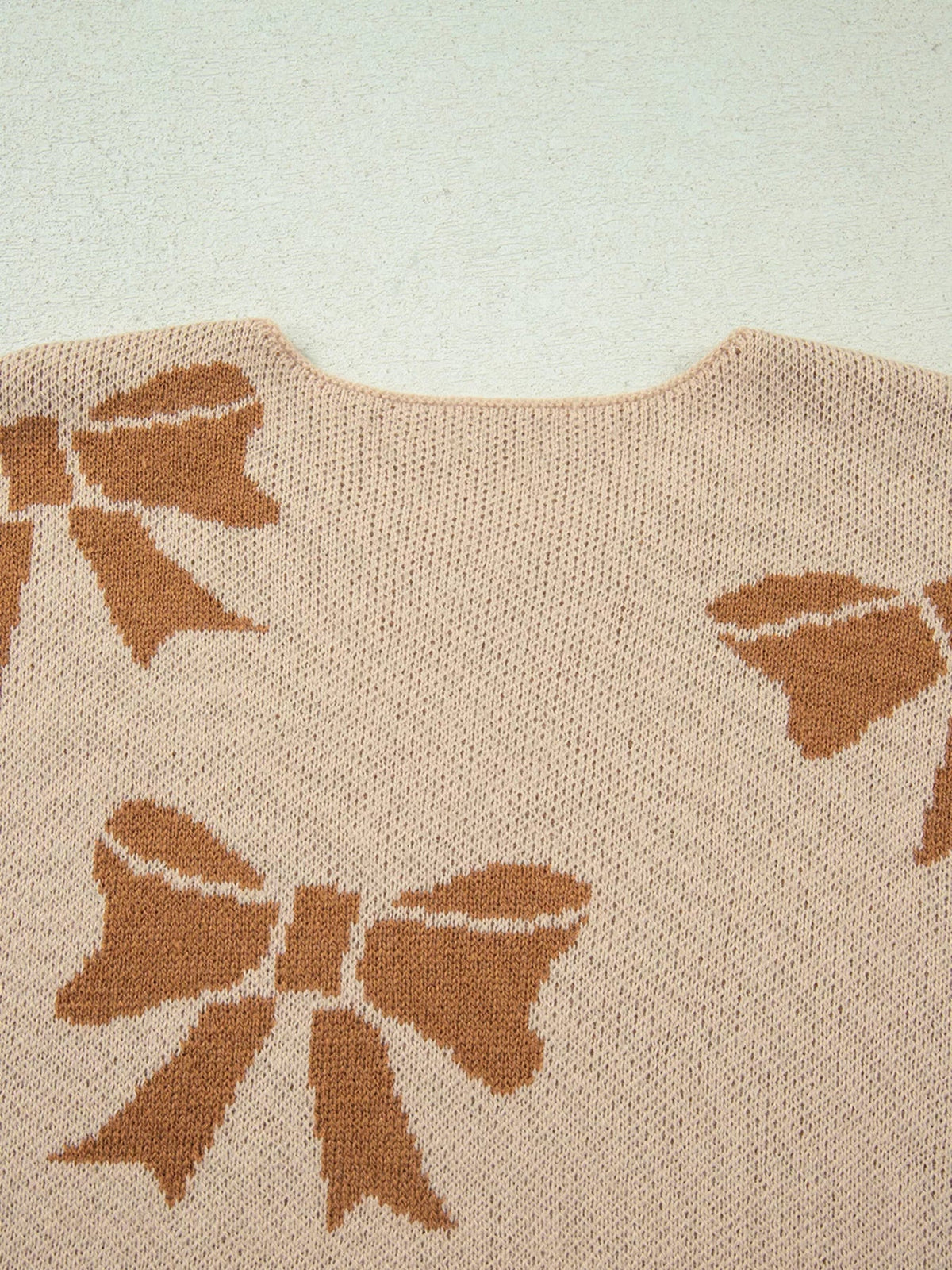 Close-up of a beige knit sweater with brown ribbon bow motifs, a round neckline, and textured construction.