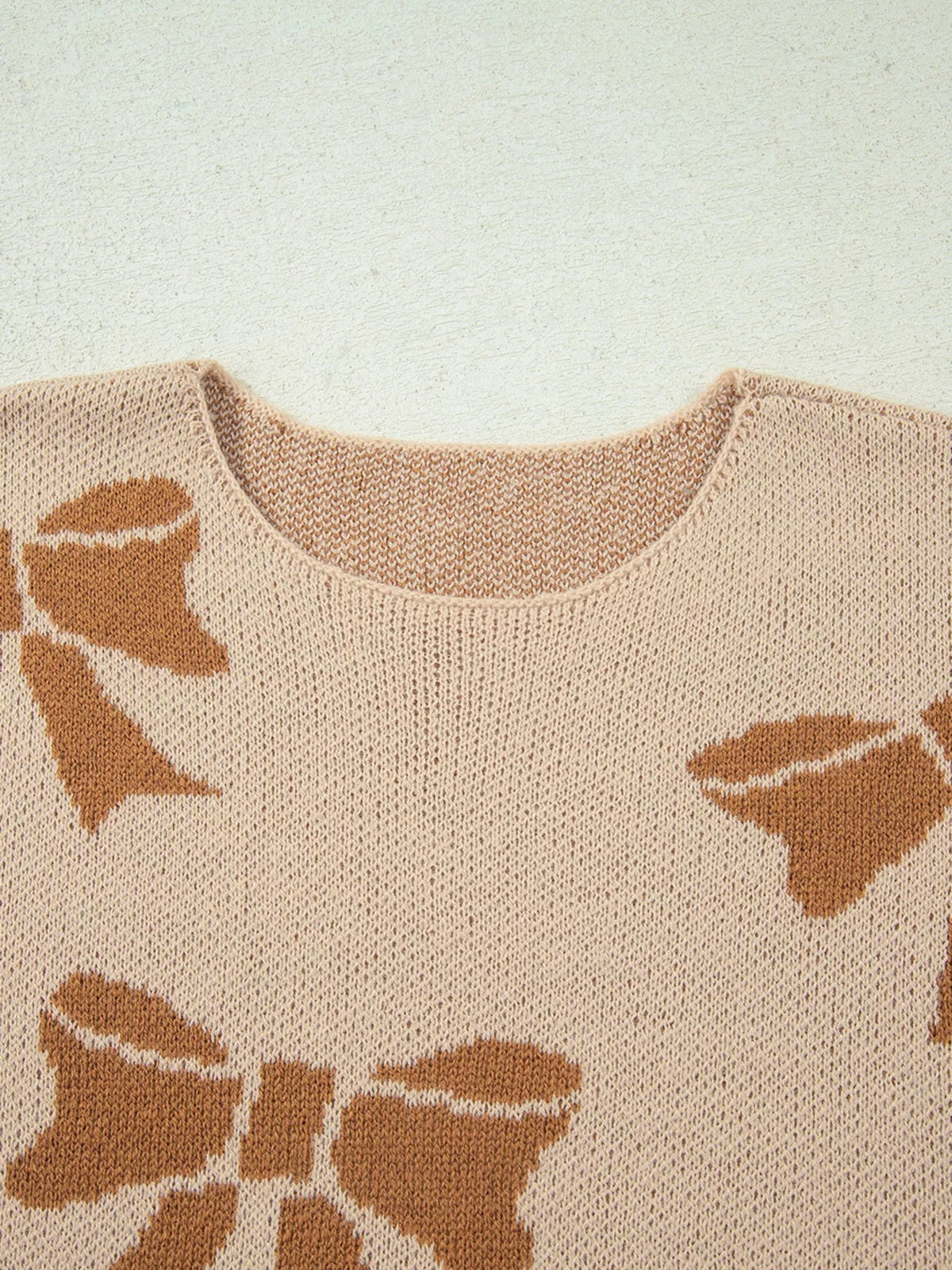 Close-up of a cream knit sweater with a rounded neckline, brown ribbon bow motifs, and visible textured stitching across the front.