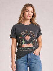 Model wearing a charcoal graphic round-neck short-sleeve T-shirt with rolled cuffs, relaxed silhouette, and colorful “RISE WITH THE SUN” desert print.