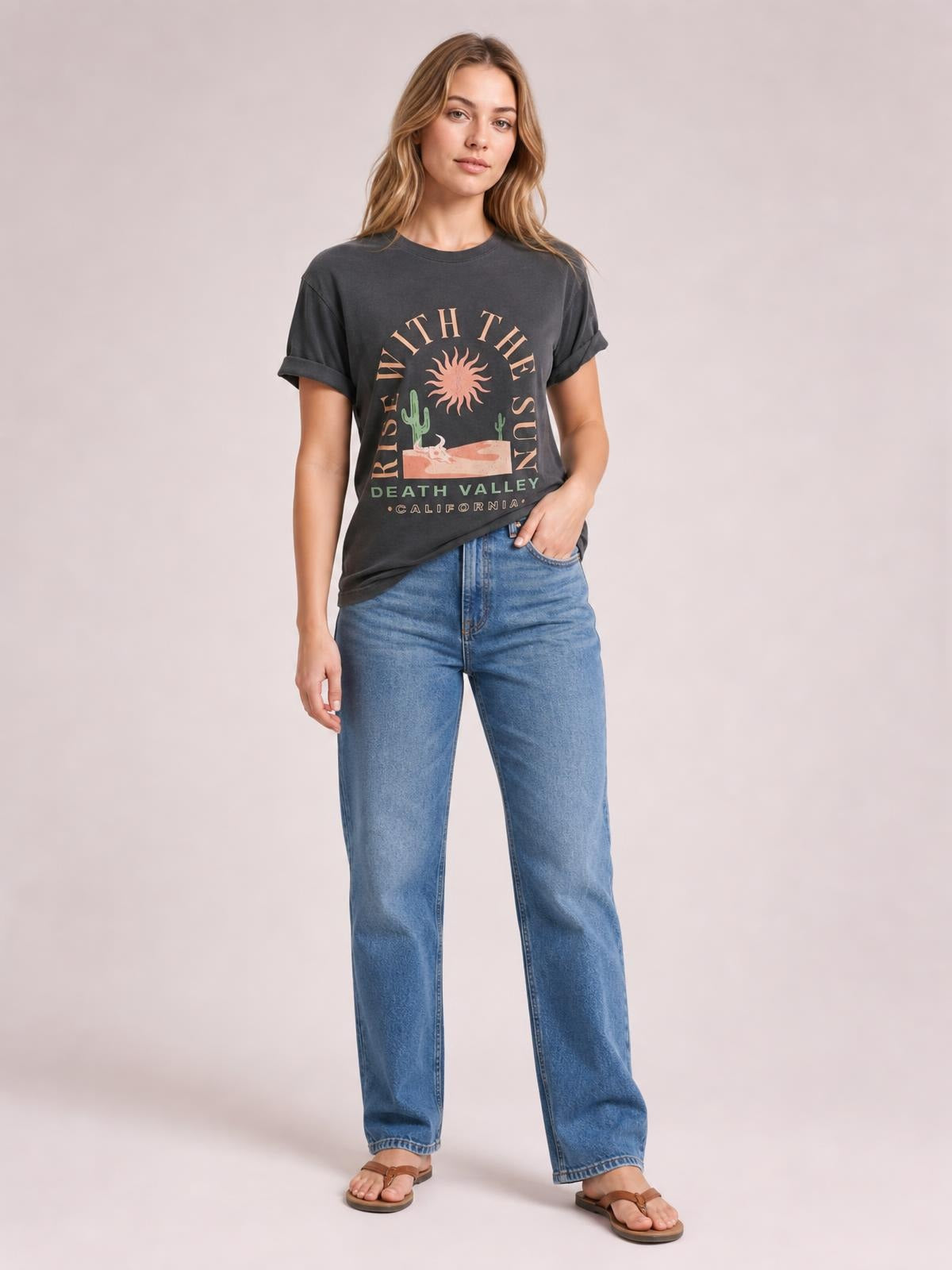 Model wears a charcoal RISE WITH THE SUN graphic T-shirt with a round neck, short sleeves, rolled cuffs, and a relaxed silhouette, posing with one hand in her jeans pocket.