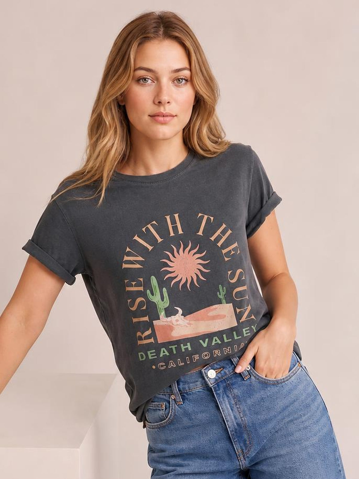 A model wears a charcoal RISE WITH THE SUN graphic T-shirt with a round neck, rolled short sleeves, relaxed cropped silhouette, and one hand in her jeans pocket.