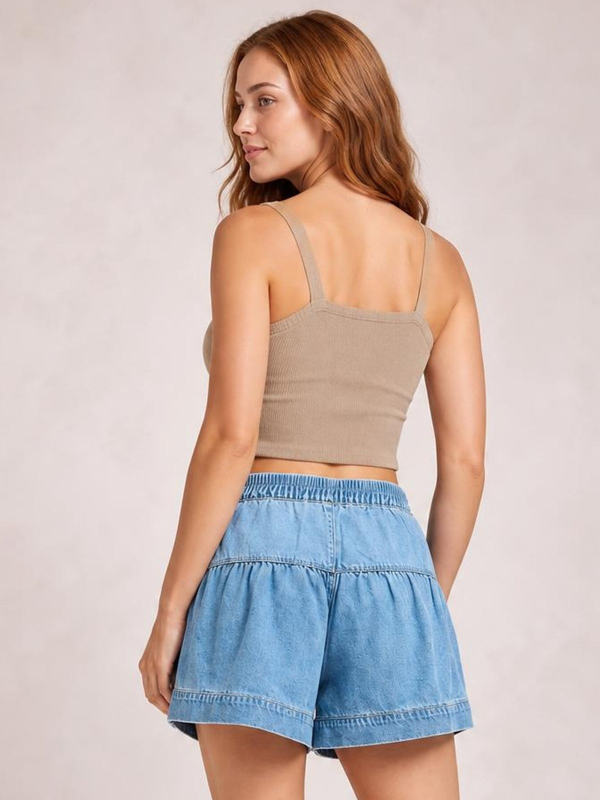 A model poses from behind wearing light blue slouchy shorts with an elastic shirred waistband, loose silhouette, and stitched tiered construction.