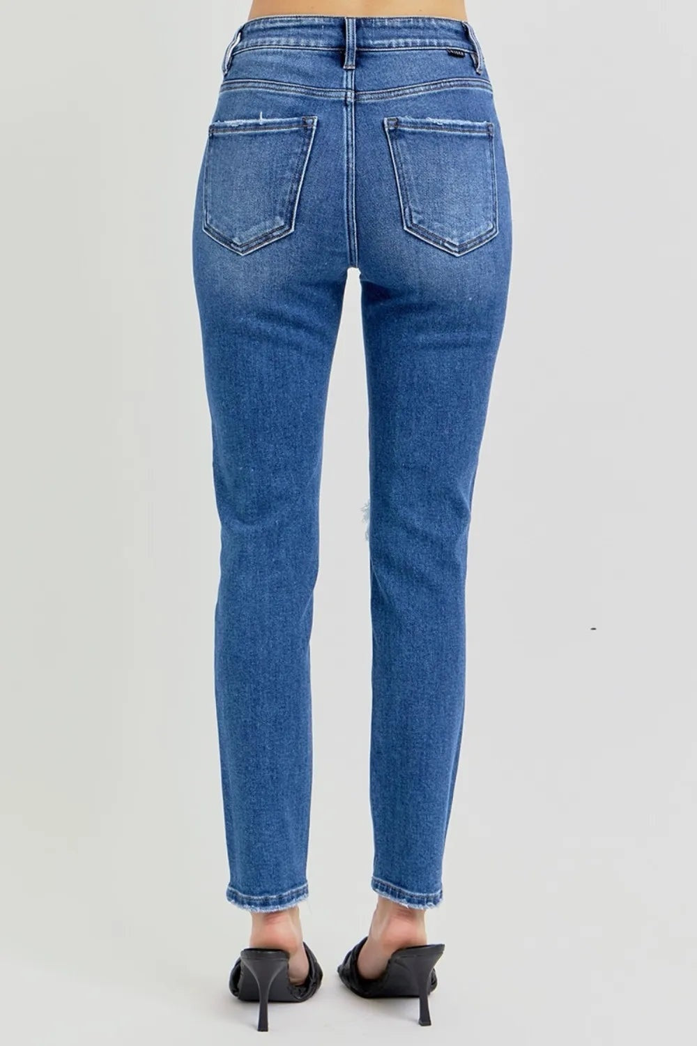 Rear view of a model wearing blue washed high-rise ankle skinny jeans with knee distressing, belt loops, and two outlined back pockets.