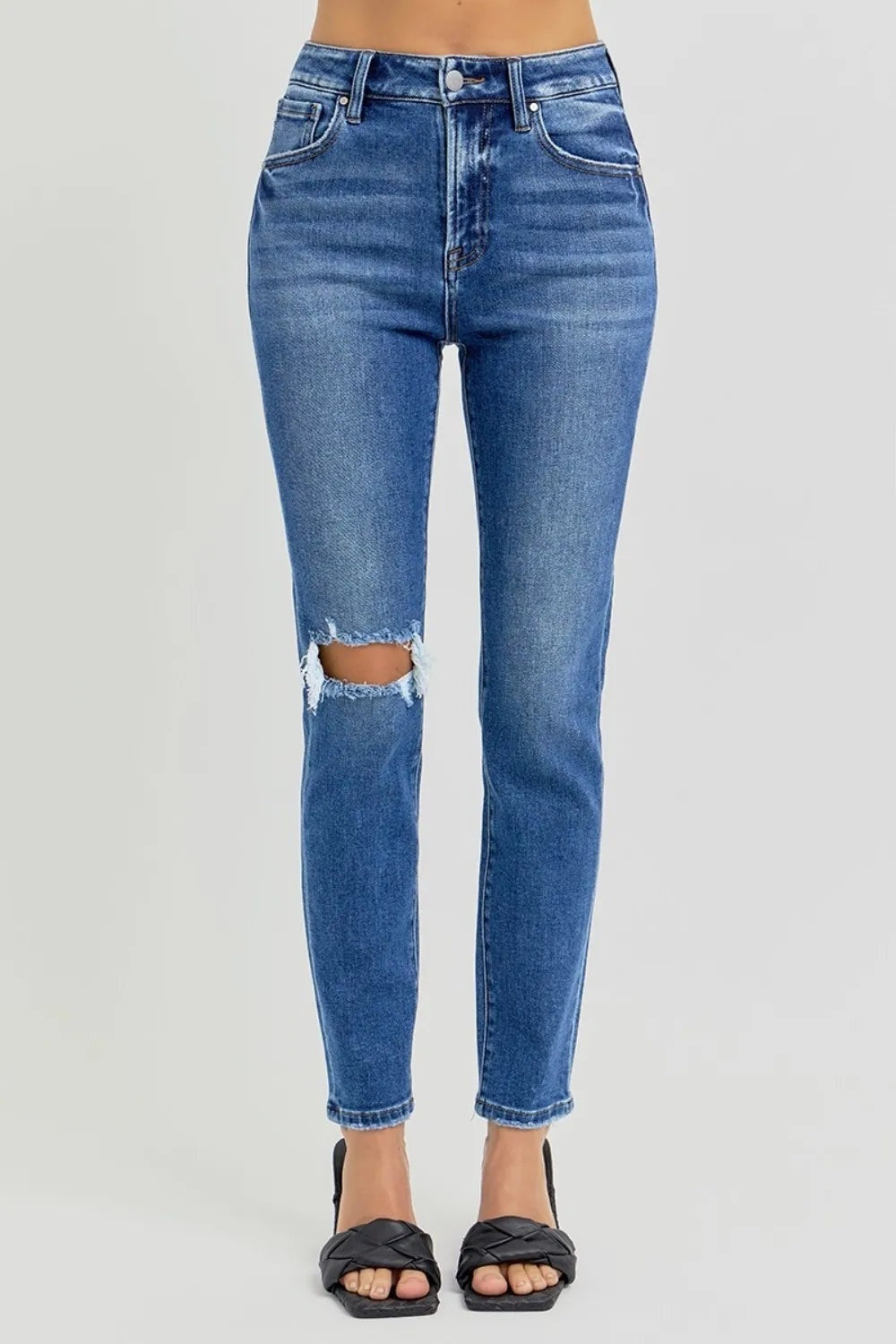 A standing model wears washed blue high-rise ankle skinny jeans with front pockets, a fitted silhouette, frayed knee distressing, and cropped hems.