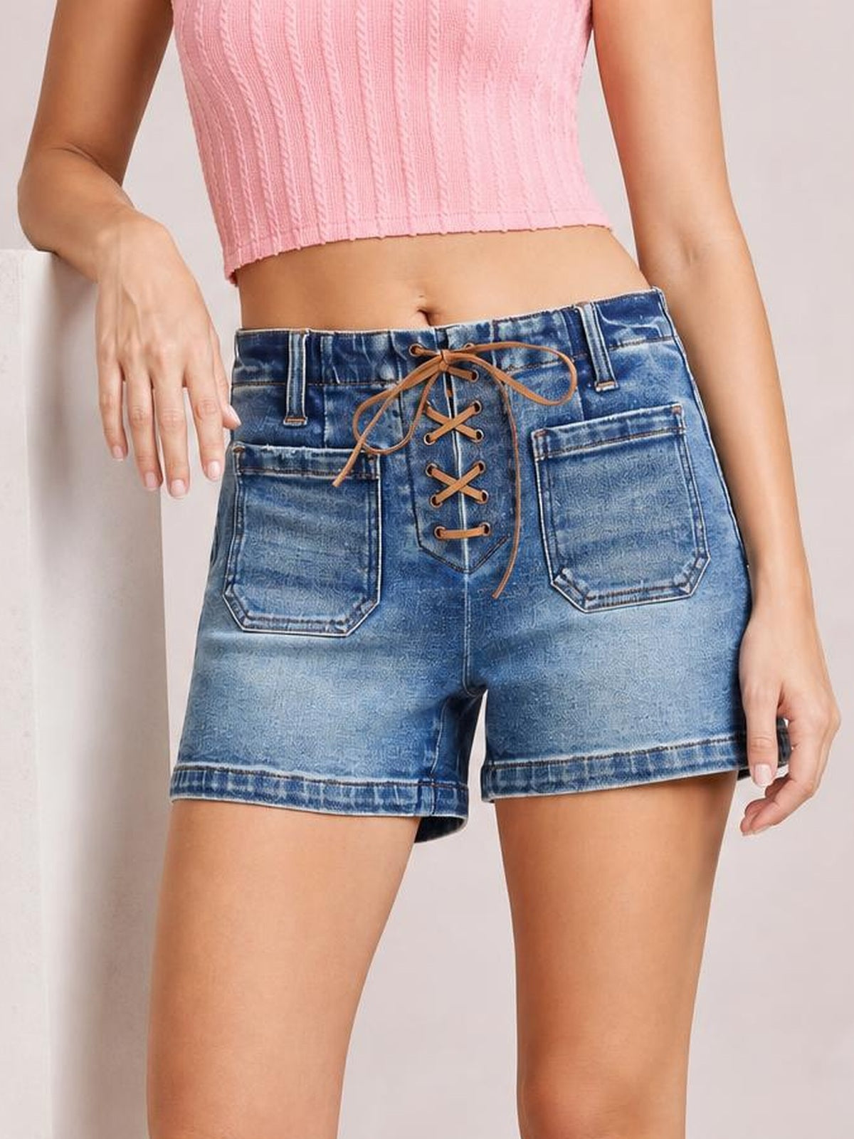 Model wears blue high-rise denim shorts with a relaxed silhouette, front lace-up tie detail, belt loops, stitched hems, and large patch pockets.
