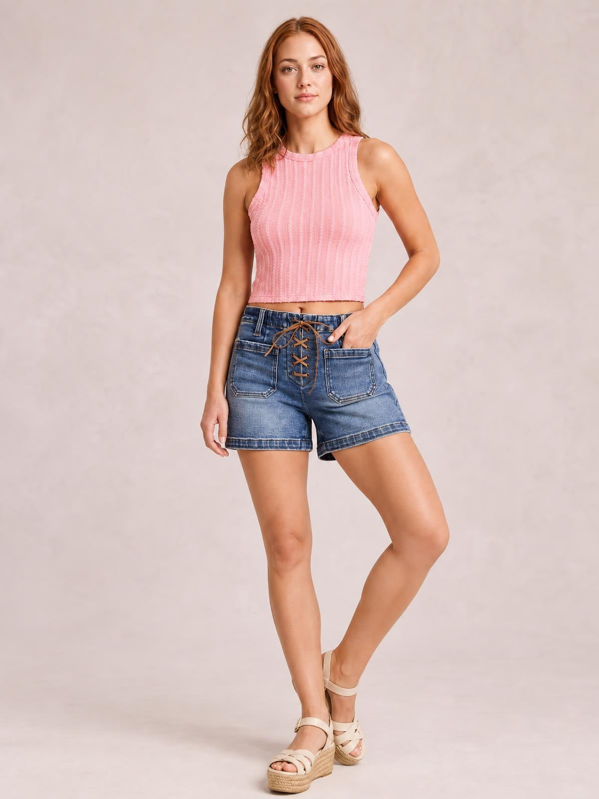 Model poses in high-rise blue denim shorts with a front tie-up lace detail, patch pockets, relaxed silhouette, and cuffed hems.
