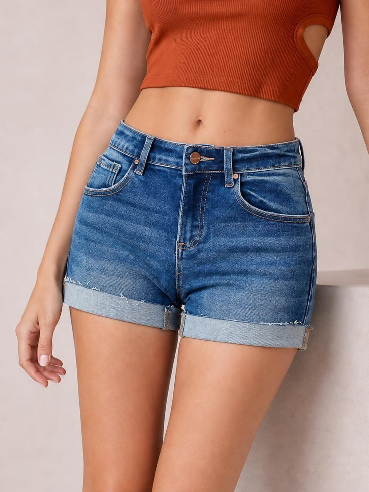 A model stands in low-rise blue denim cuffed shorts with a relaxed silhouette, five-pocket construction, belt loops, front button, and raw-edged rolled hems.