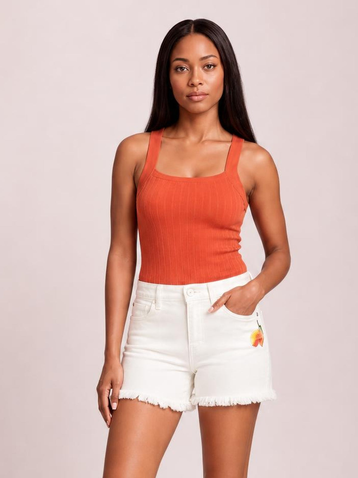 A model wears white high-rise shorts with a relaxed tailored silhouette, button fly, front pockets, small flower embroidery, and raw hems, posing with one hand in a pocket.
