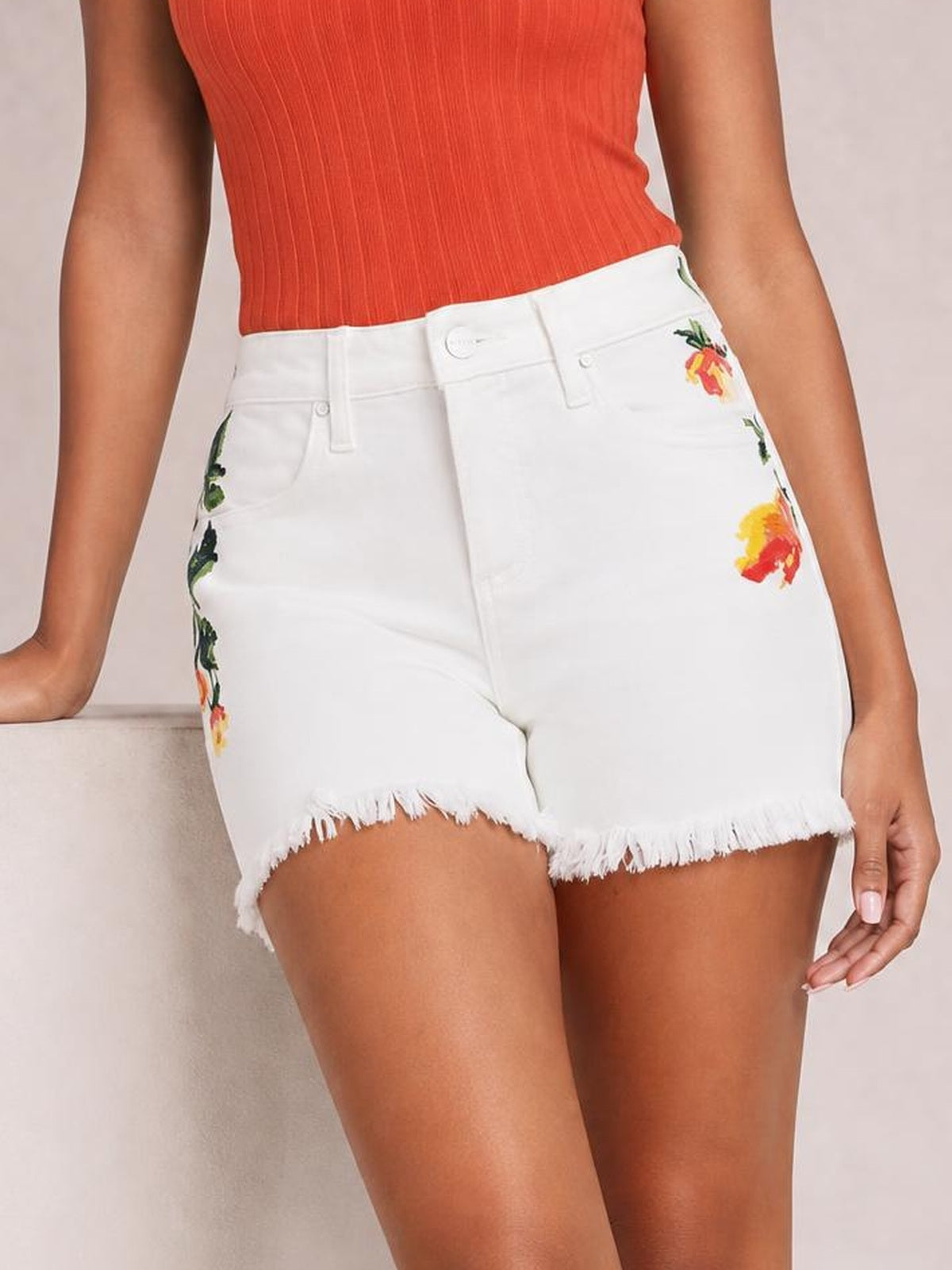 A model wears white high-rise tummy-control shorts with colorful embroidered flowers, front pockets, a structured fit, and a raw frayed hem.