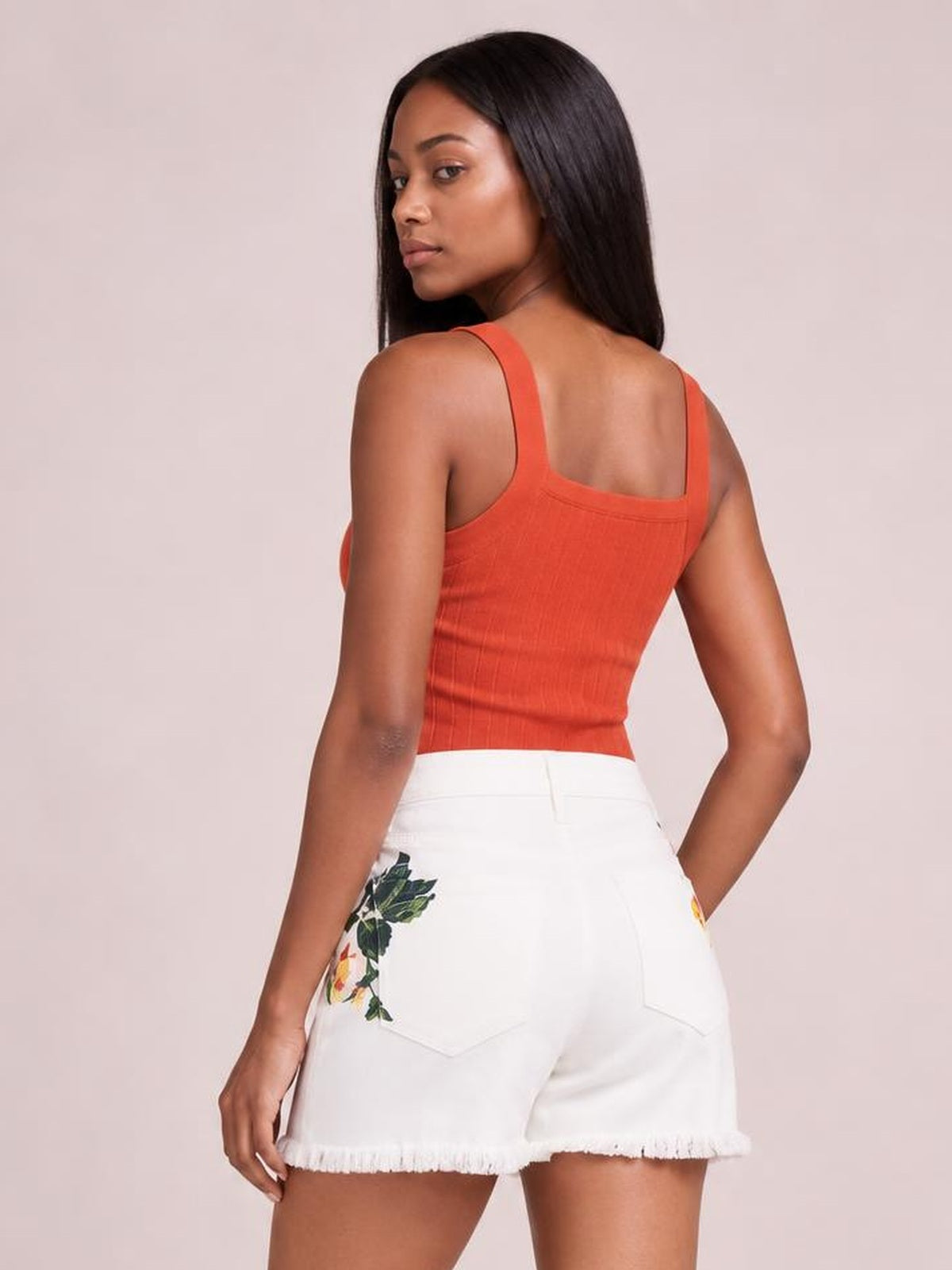 A model poses in a rear three-quarter view wearing white high-rise flower-printed shorts with embroidered details, back pockets, and a relaxed raw hem.