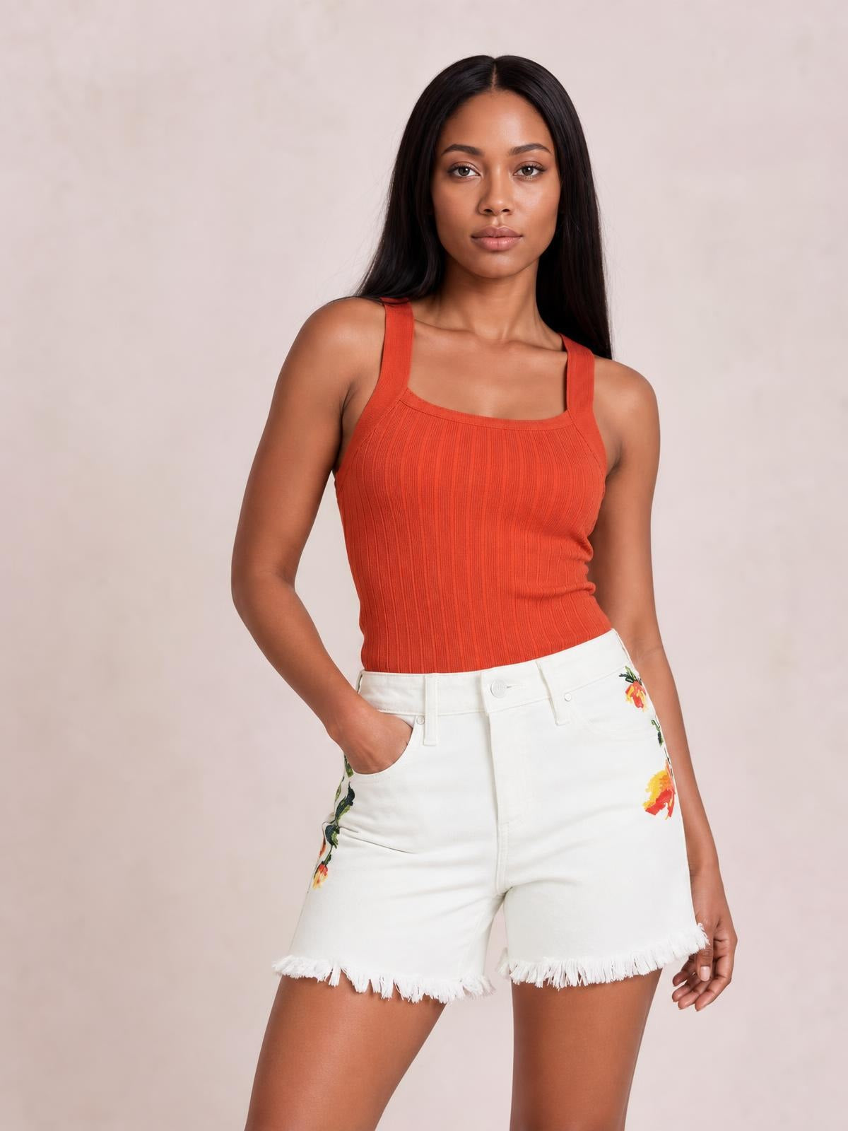 A model poses in white high-rise flower-embroidered shorts with a relaxed tailored silhouette, tummy control waistband, front pockets, and raw fringed hems.