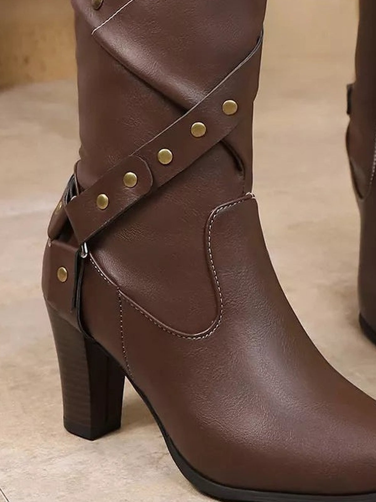 Close-up of brown PU leather heeled boots featuring criss-cross riveted straps, stitched paneling, ankle coverage, and sturdy high block heels.