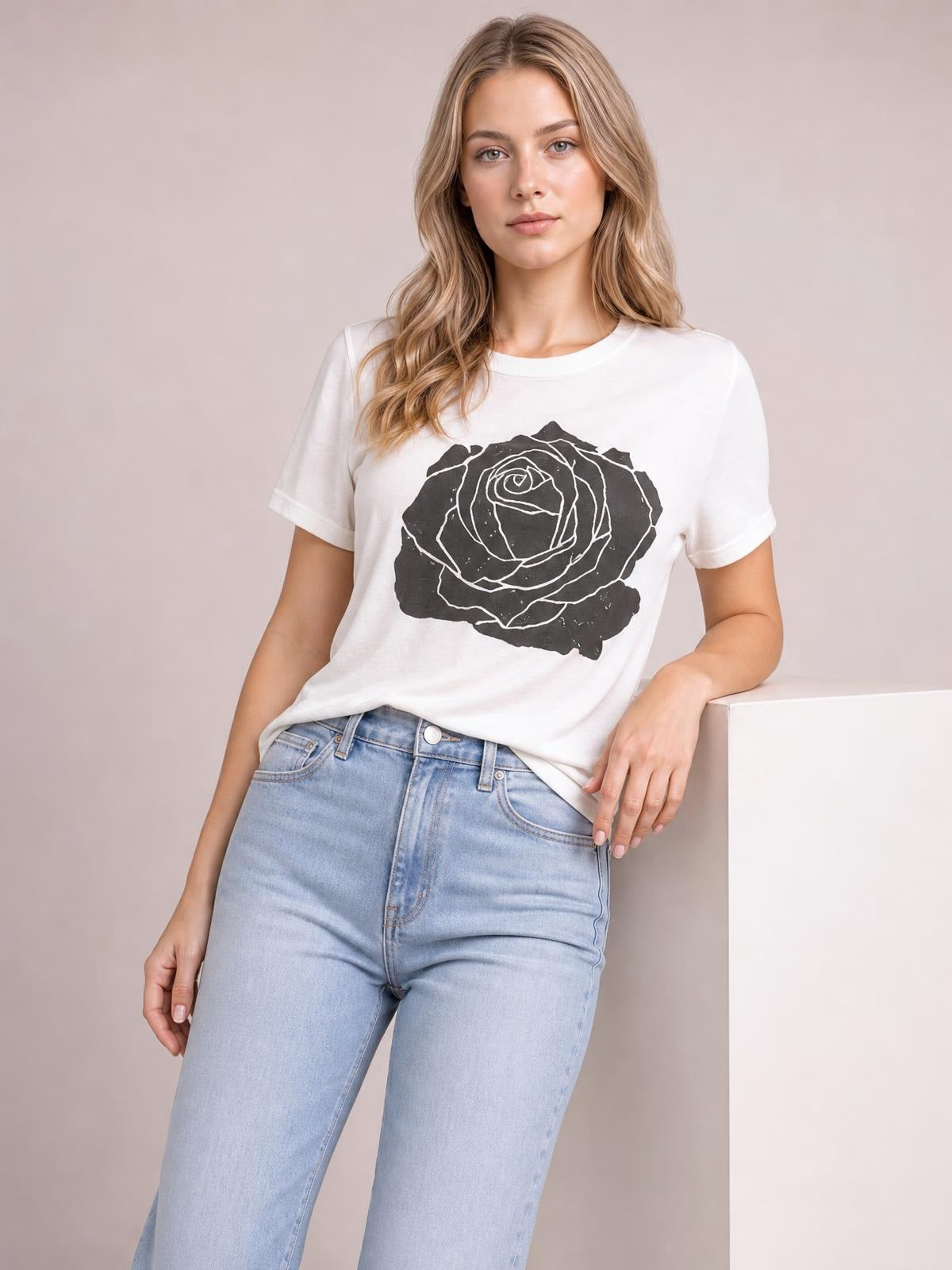 Model wears a white round-neck short-sleeve T-shirt with a bold black rose graphic, relaxed straight silhouette, and slightly tucked hem while leaning casually.