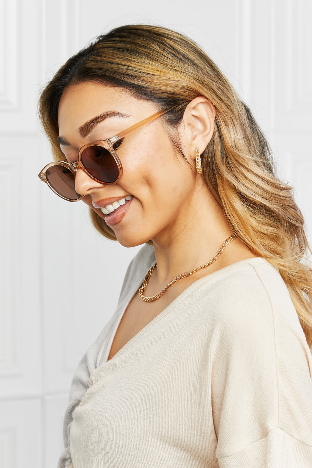 A smiling model turns in side profile wearing round full-rim sunglasses with translucent brown polycarbonate frames and dark lenses over a cream top.
