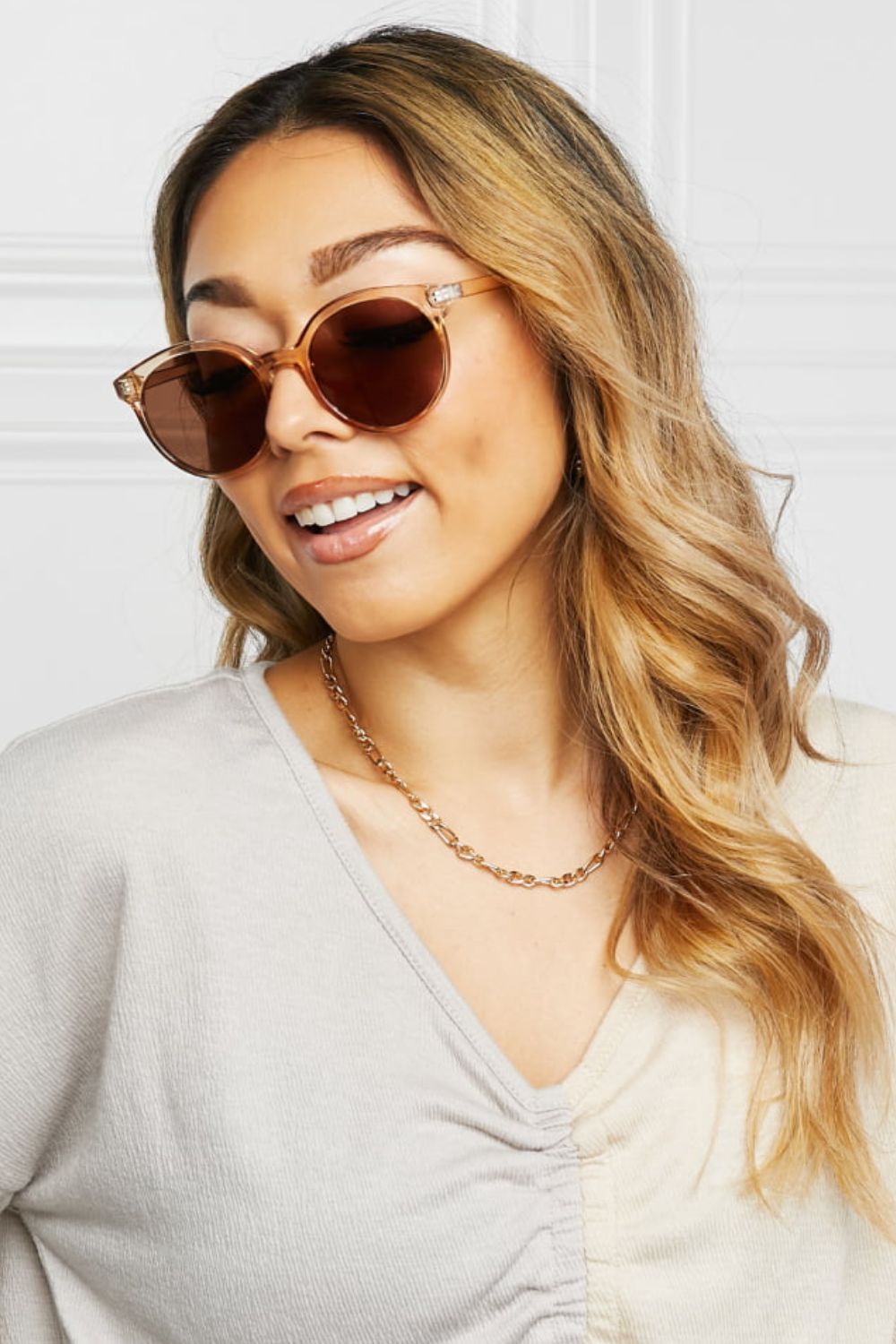 Model smiling and turning slightly wears brown round full-rim sunglasses with softly curved lenses, polished polycarbonate frames, and defined temples.