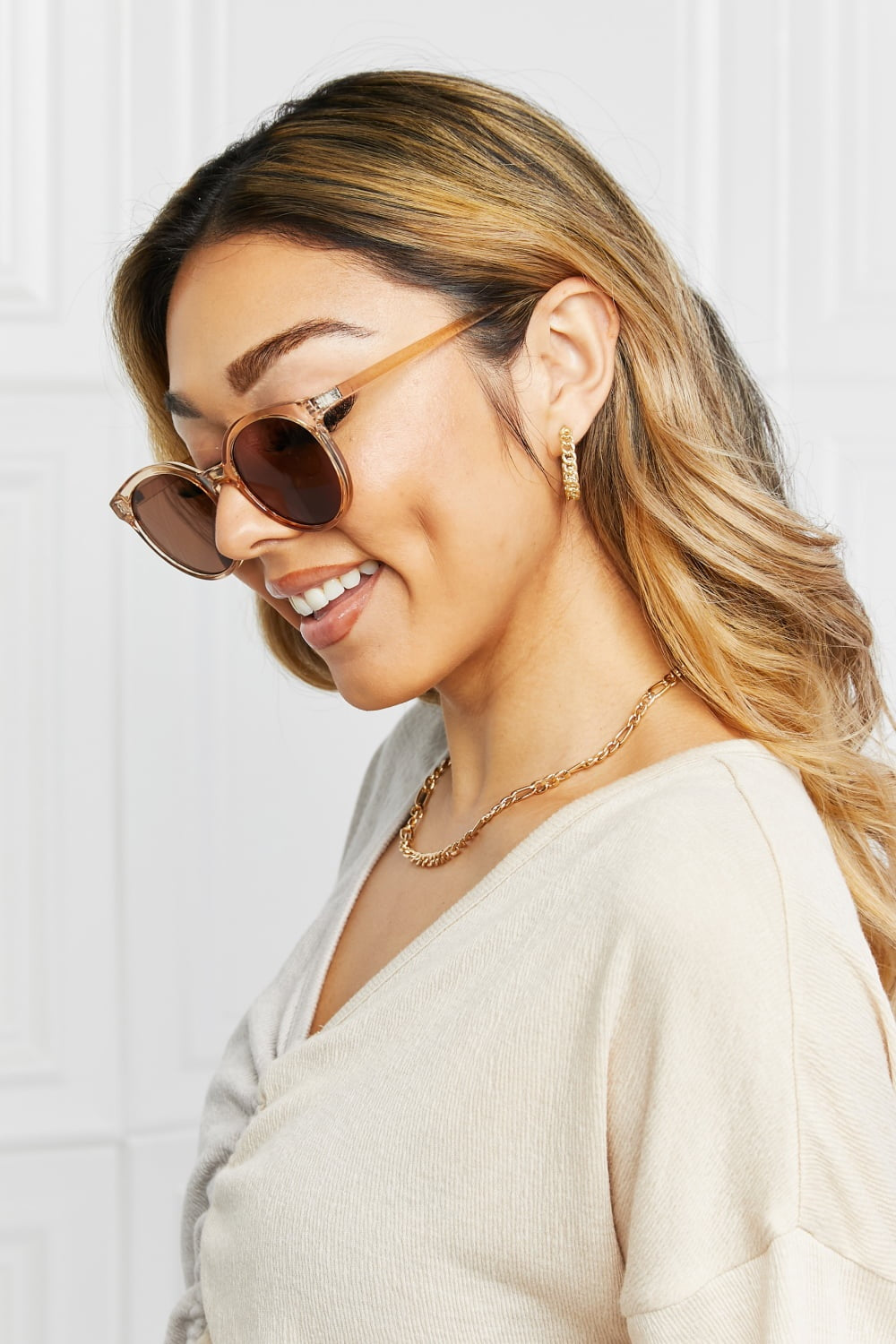 A smiling model turns in side profile wearing round full-rim sunglasses with translucent brown polycarbonate frames and dark lenses over a cream top.