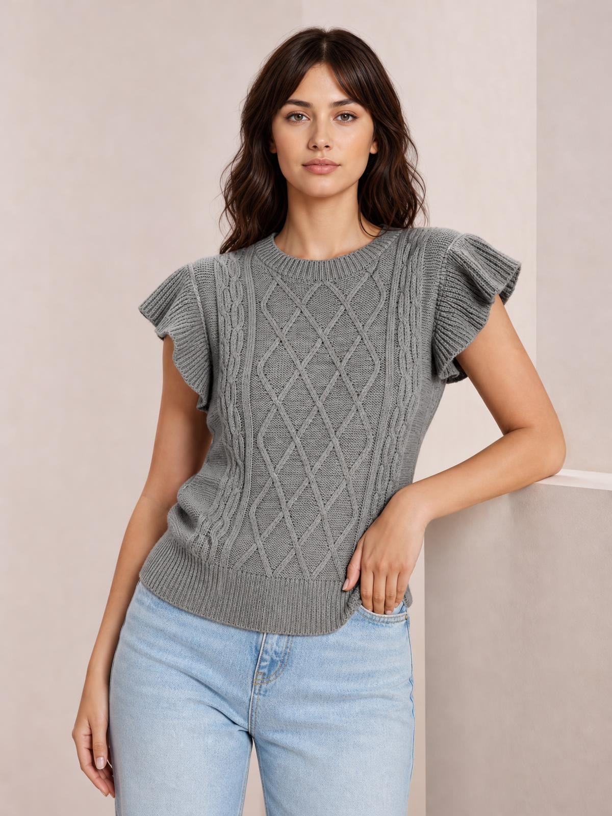 Model wearing a gray round neck cable-knit sweater with ruffled cap sleeves, a fitted silhouette, and ribbed neckline and hem, posing with one hand in her jeans pocket.