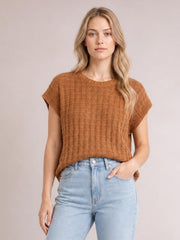 Model wears a brown round-neck sweater vest with cap sleeves, textured knit construction, and a relaxed straight silhouette, posing with one hand in her jeans pocket.