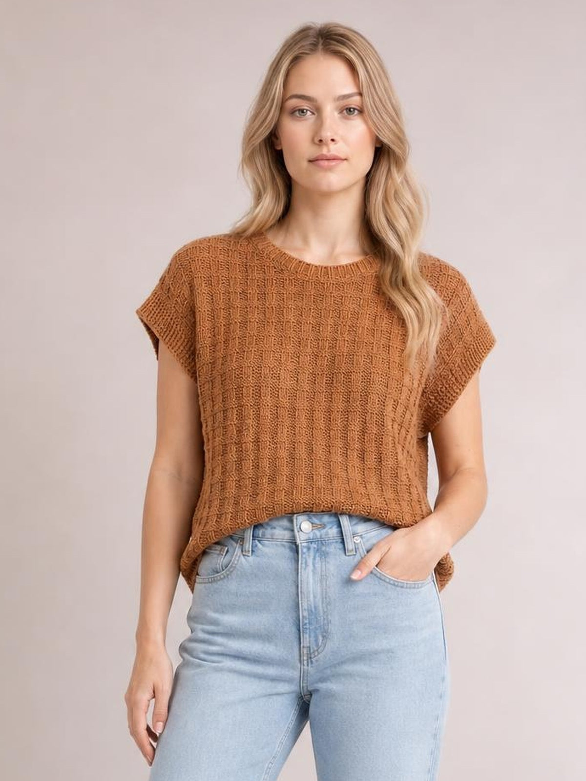 Model wears a brown round-neck sweater vest with cap sleeves, textured knit construction, and a relaxed straight silhouette, posing with one hand in her jeans pocket.
