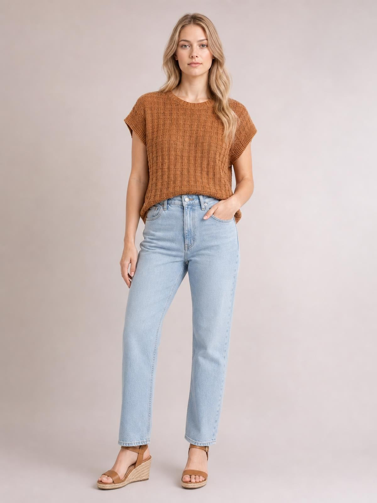 Model wears a rust-brown round neck cap sleeve sweater vest with textured knit construction and a relaxed, slightly boxy silhouette, posing with one hand in her jeans pocket.