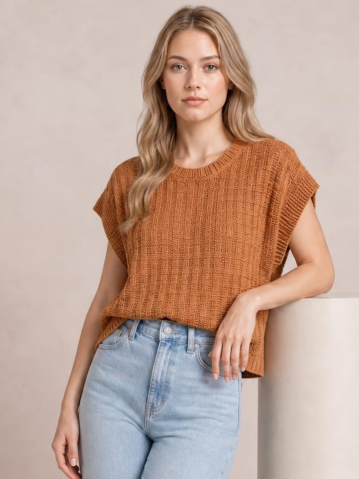 Model wears a rust-brown round neck cap sleeve sweater vest with textured knit construction and a relaxed silhouette, posing with one hand resting at her hip.