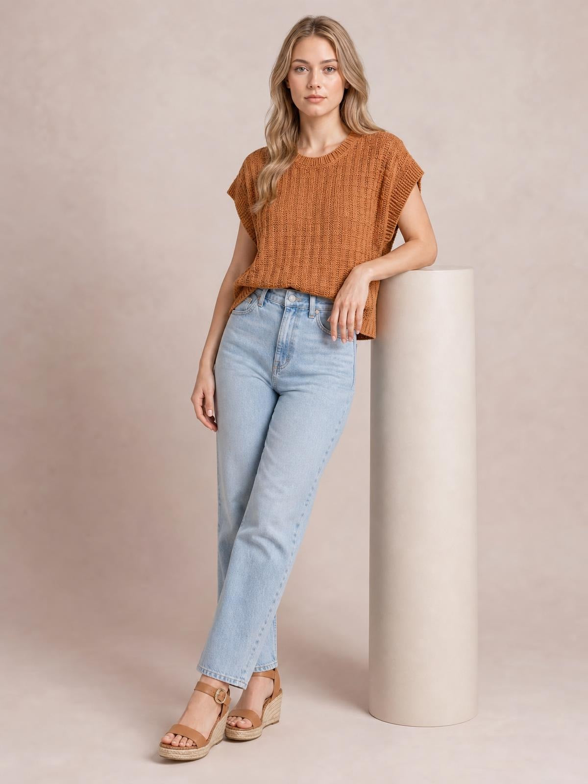 A model leans against a column wearing a rust-brown round neck cap sleeve sweater vest with a relaxed silhouette and textured knit construction.