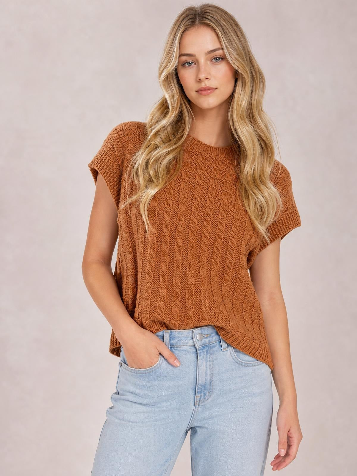 Model wears a rust-brown round neck cap sleeve sweater vest with textured knit construction and a relaxed, straight silhouette, paired with light-wash jeans.